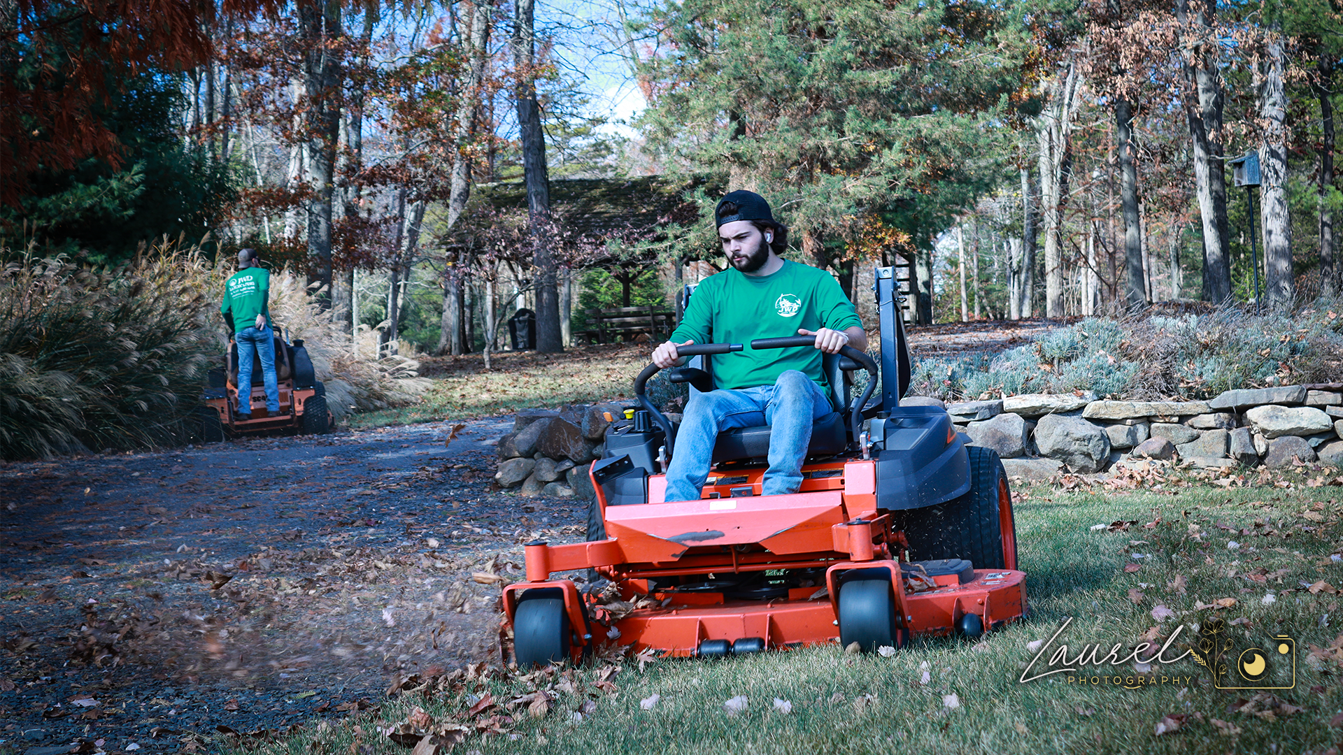 Two men working outdoors in a wooded area during autumn, one man is sitting on a riding lawn mower and the other man is standing in the background near a lawn tractor, surrounded by trees with fall foliage.