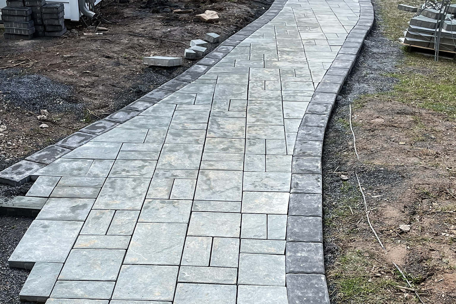 Newly paved concrete walkway with a curved design, bordered by darker concrete stones, in an outdoor setting with bare soil and construction materials nearby.