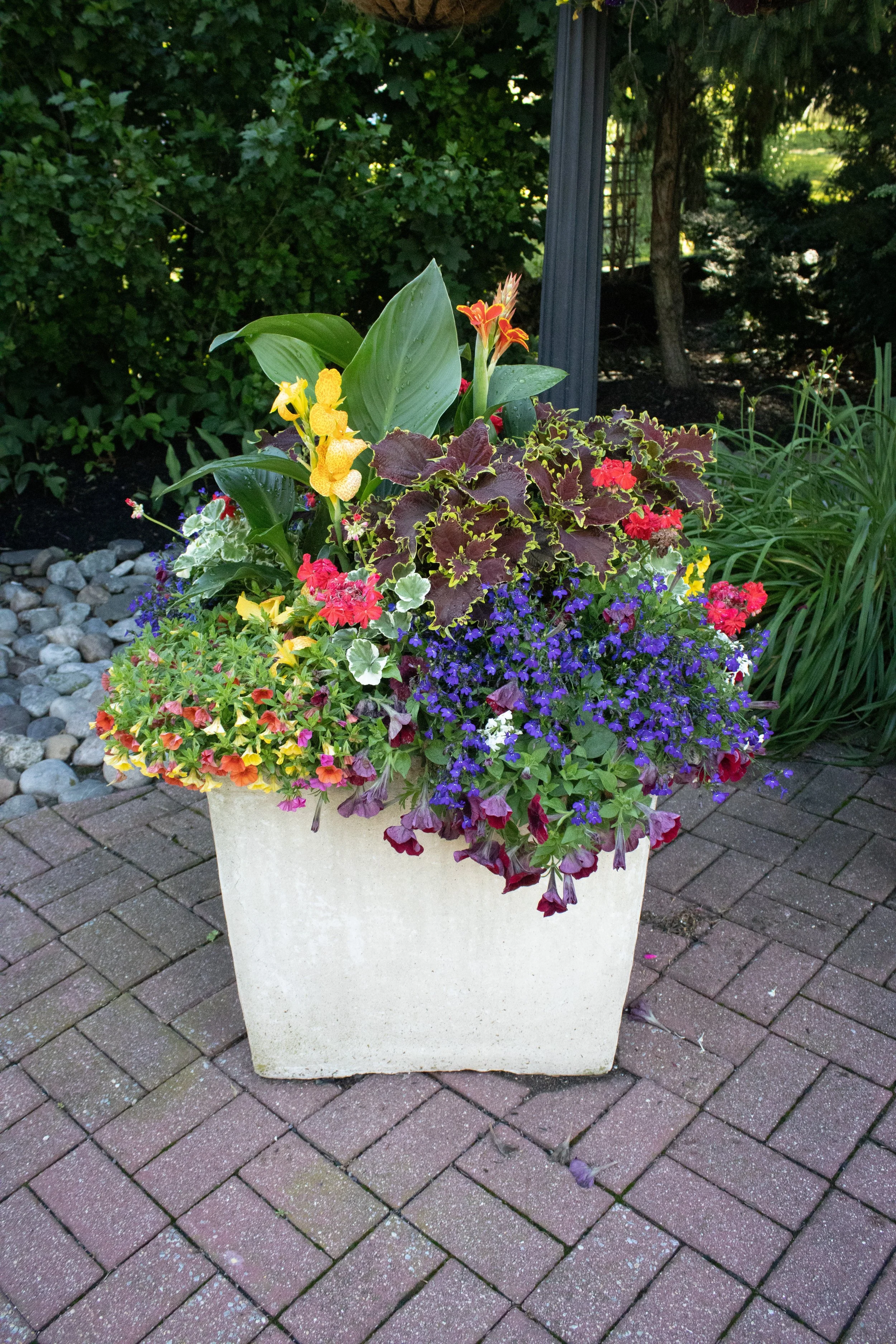 A large beige planter with various colorful flowers and green foliage, placed on a brick-paved surface outdoors.