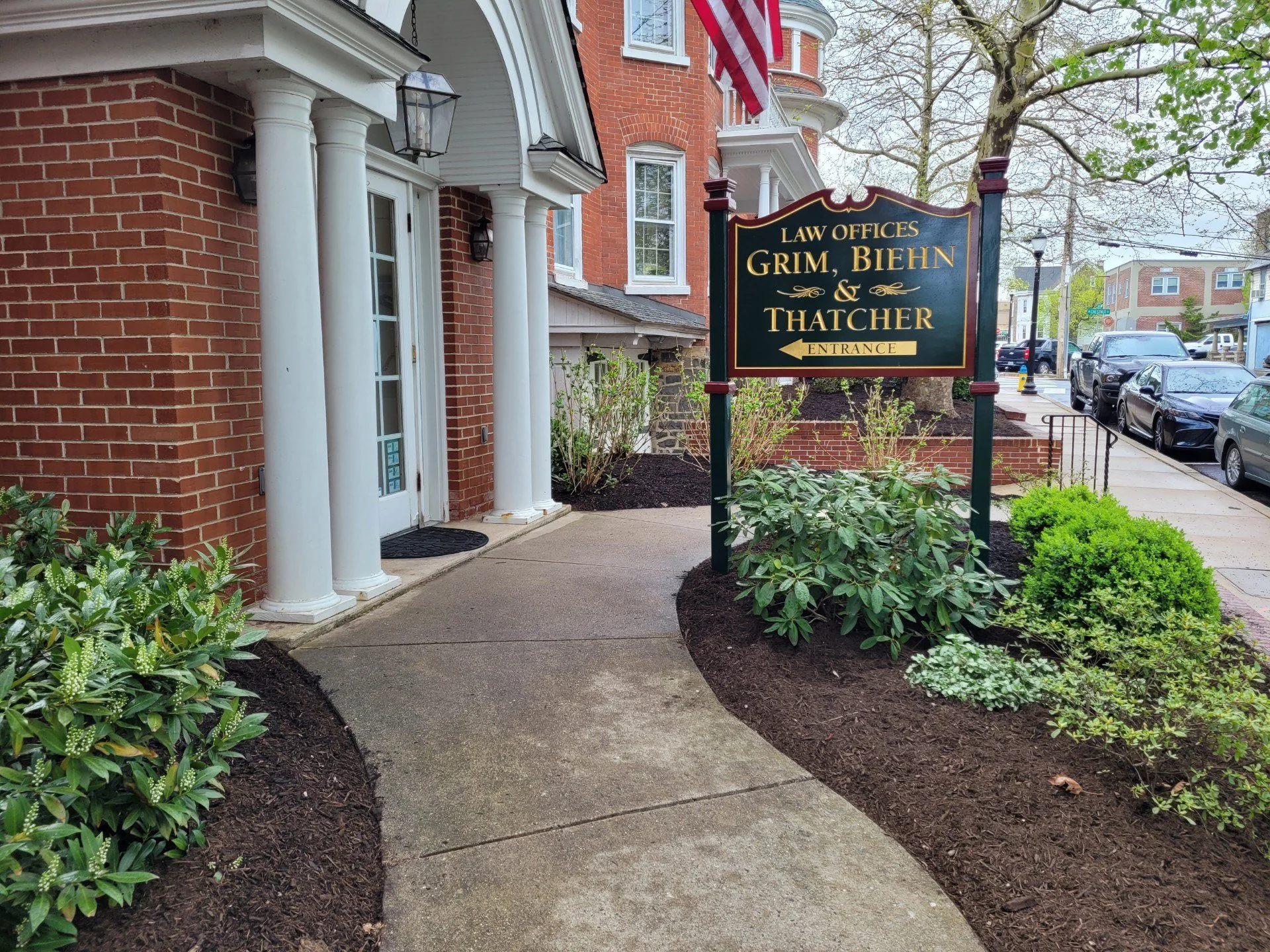 Sidewalk leading to law office building with columns, shrubbery, and a sign reading 'Law Offices Grim, Biehn & Thatcher' near the entrance, with parked cars and trees in the background.