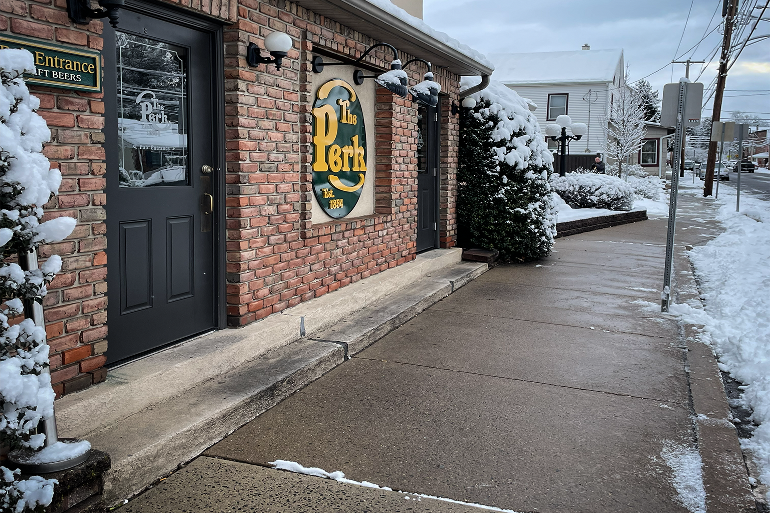 Snow-covered sidewalk outside a brick building with a sign that reads 'The Perk.' Snow is on trees and shrubs, with cloudy sky overhead.