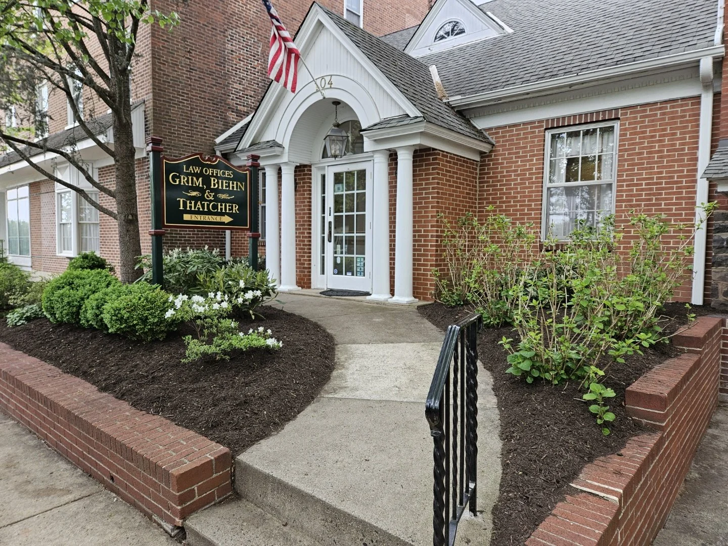 The entrance of a building with a sign for law offices of Grim, Biehn, and Thatcher. The building has brick walls, a white door, and a small porch covered by a gabled roof. There are well-maintained shrubs and flowers along the sidewalk, and an American flag is mounted above the sign.