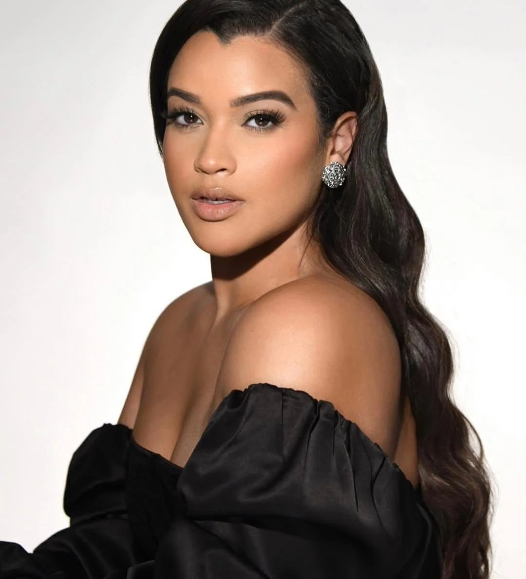 A woman with long wavy dark hair, Hollywood waves, wearing a black off-shoulder top with puffed sleeves and large diamond earrings, looking at the camera against a plain light background.