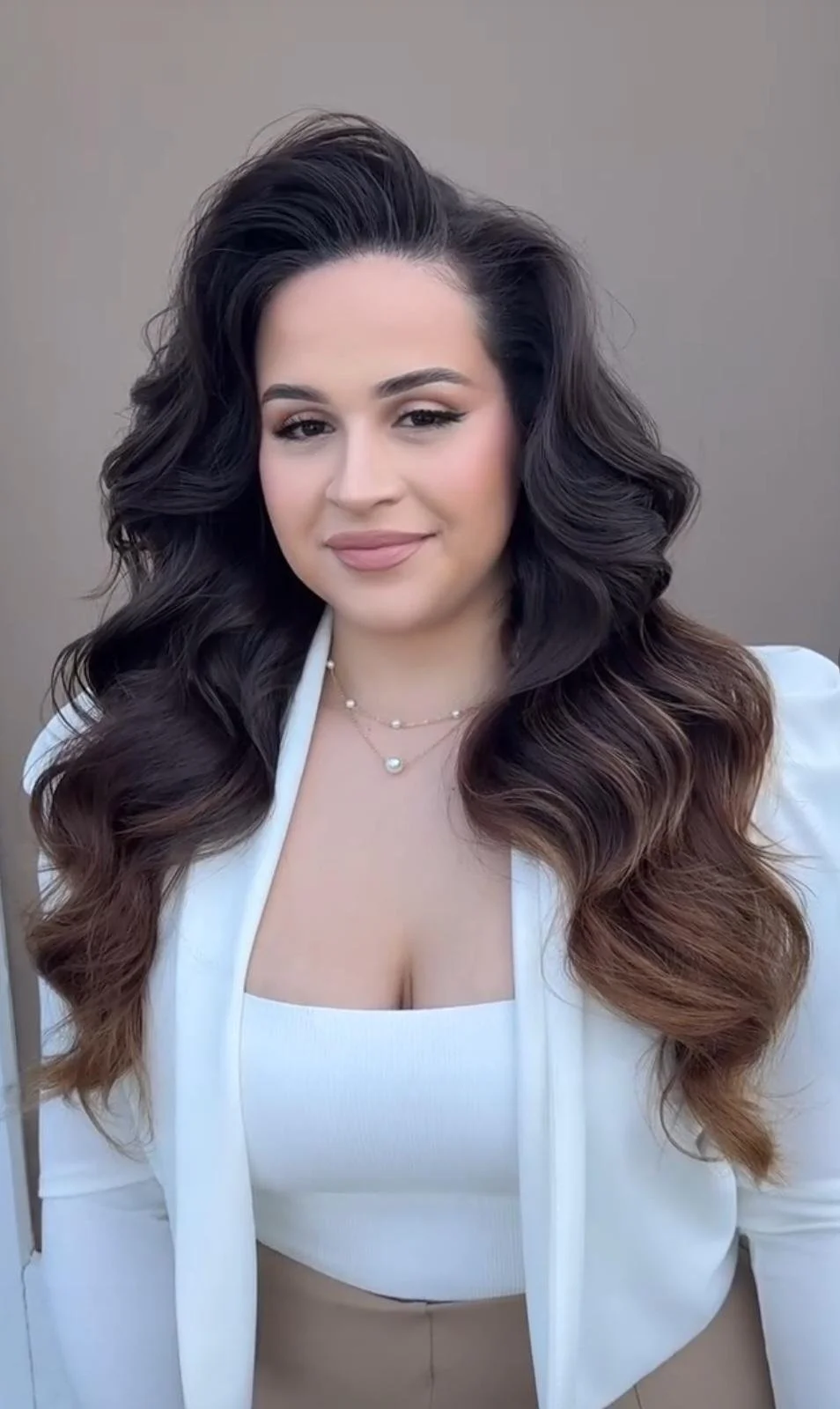 A woman with long, wavy brown hair, wearing a white blazer over a white crop top, and layered pearl necklaces, standing against a neutral background.