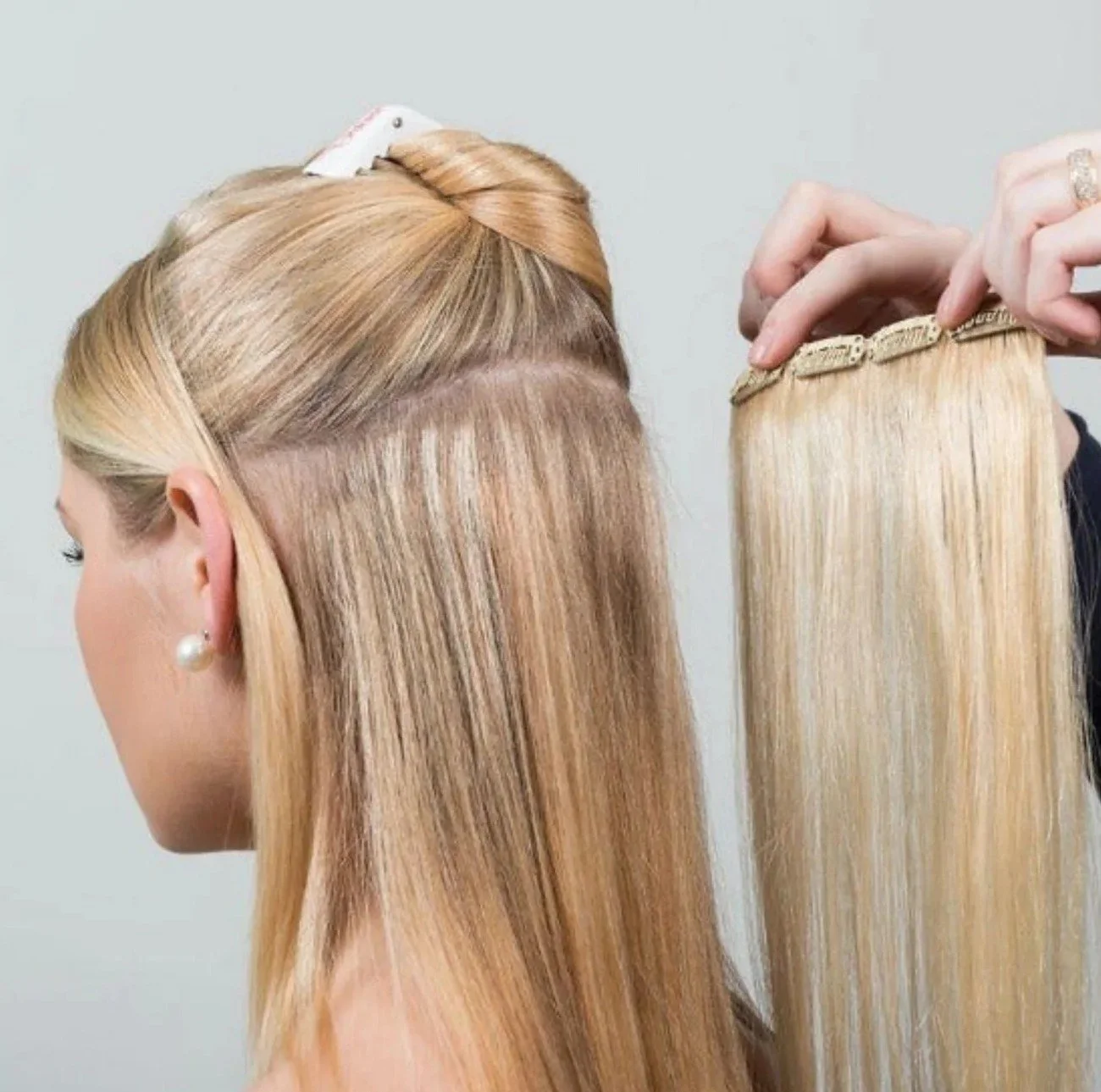 Close-up of a woman getting hair extension installation. Her blonde hair is sectioned and clamped, with a hand holding a blonde extension weft ready to be attached.