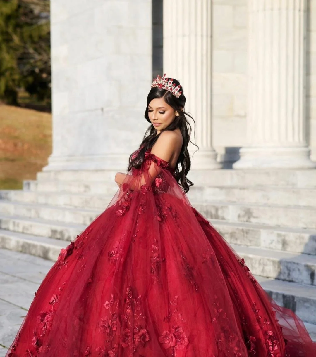 A young woman in a red ball gown with floral embellishments, standing on stone steps in front of a white marble building, wearing a matching crown, with long dark hair and a serene expression.