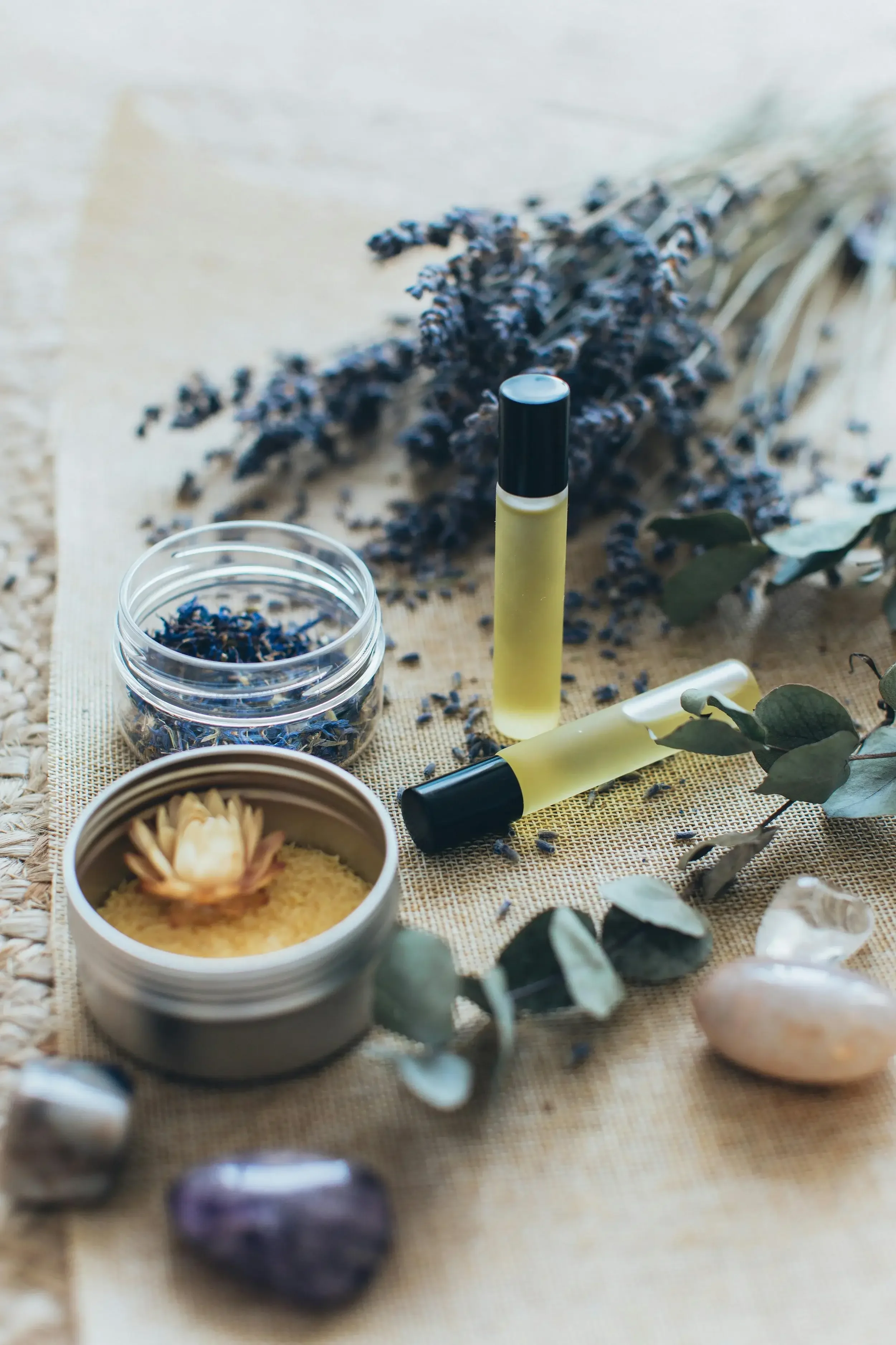 Collection of aromatherapy items including dried lavender, essential oils, a small jar of bath salts, polished stones, and eucalyptus leaves arranged on a woven surface.