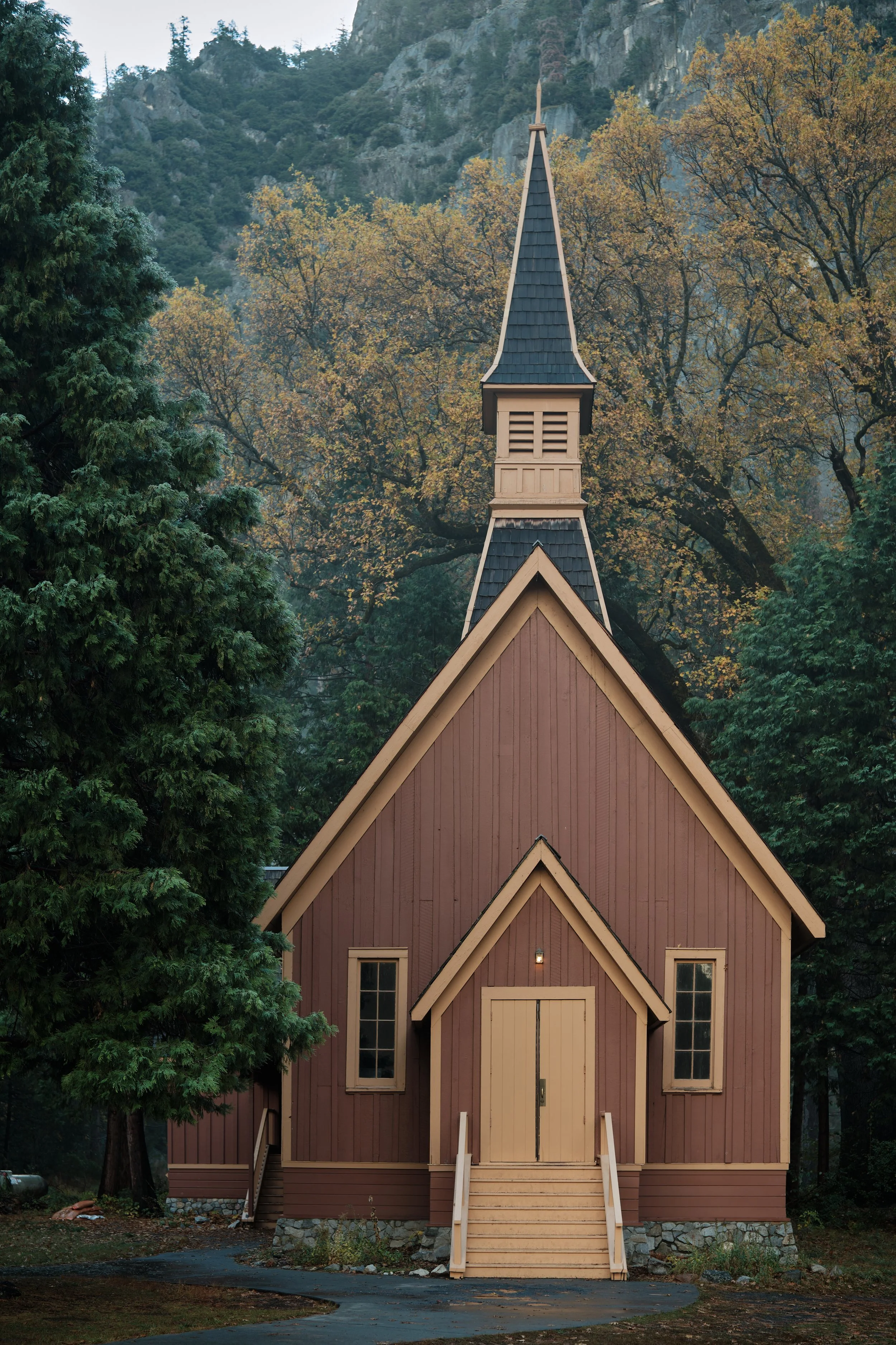 A small wooden church with a steeple, surrounded by trees with autumn foliage, set against a mountain backdrop.