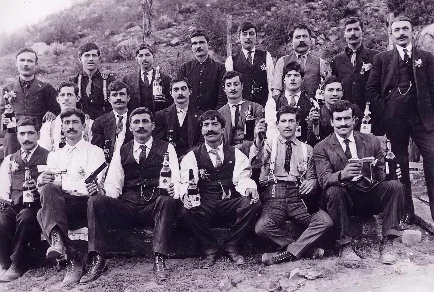 Men in their Sunday best celebrating with pistols and liquor with sprigs of vasiliko in their lapels or pockets
