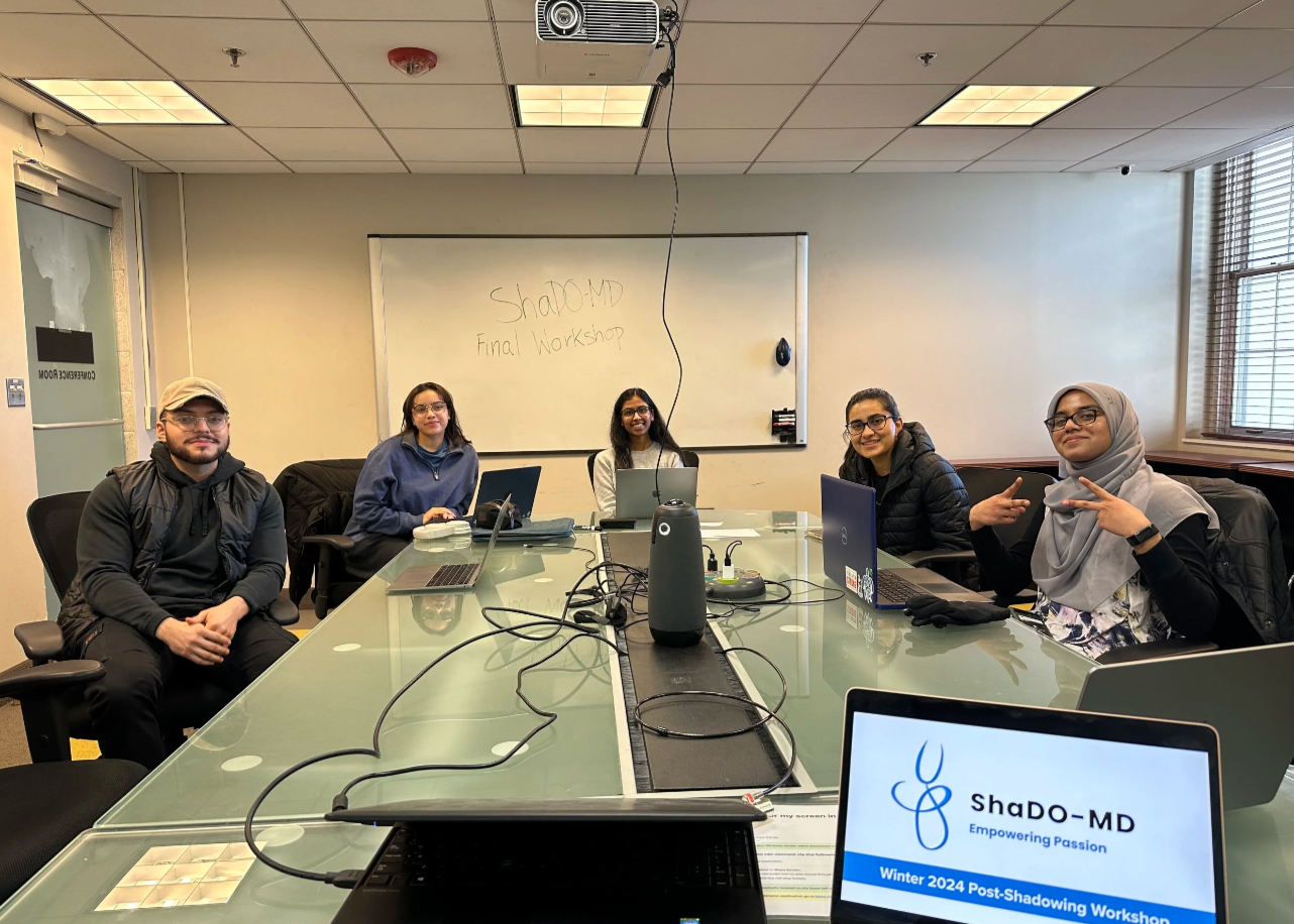 Five people seated at a conference table during a workshop, with a whiteboard in the background that reads 'ShaDO-MD Final Workshop'. There are laptops, notebooks, and a speaker on the table, and a sign on the table displaying 'ShaDO-MD Empowering Pa