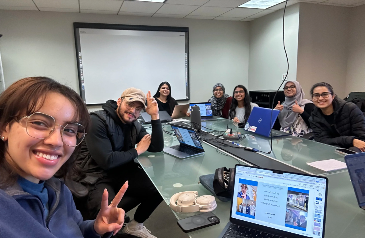 Group of seven people sitting around a conference table in a meeting room with laptops, smiling, and making peace and thumbs-up gestures. One person is taking a selfie. A large blank screen is on the wall behind them.