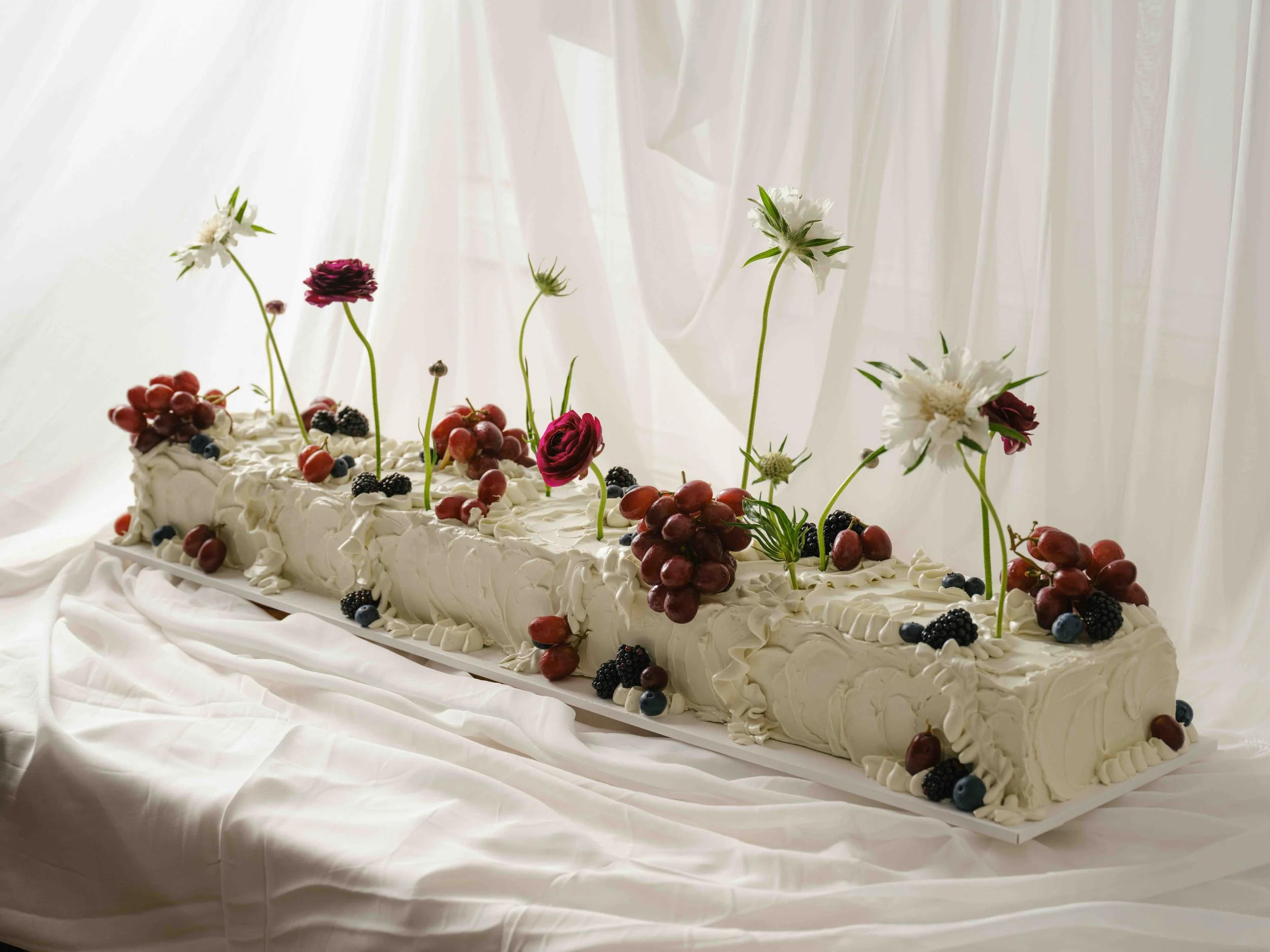 A long rectangular slab cake decorated with fresh grapes, blackberries, blueberries, and sculptural white and burgundy flowers with long green stems, set against a white fabric backdrop.