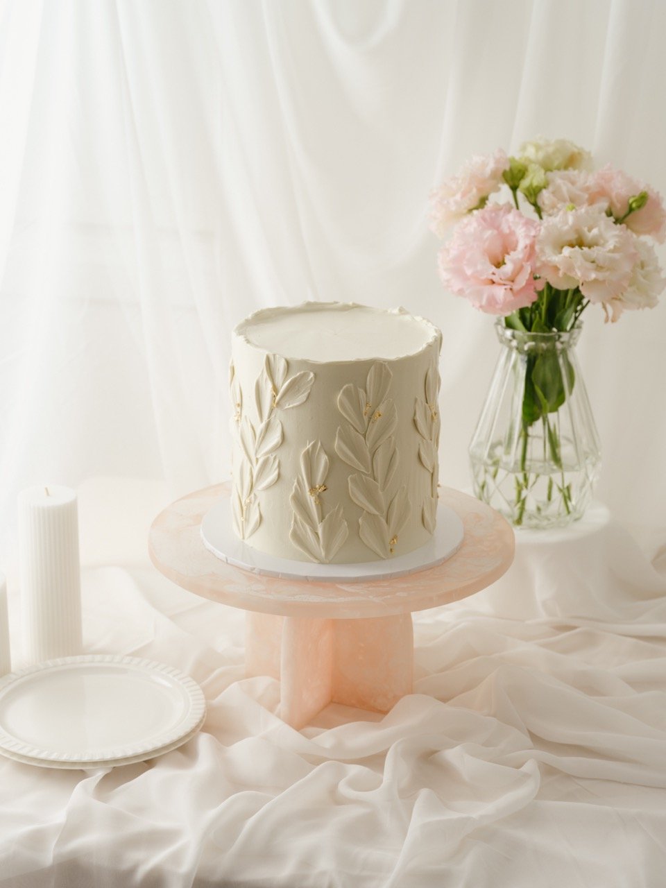 White cake with a textured buttercream tulip petal design on a pink cake stand, next to a vase of pink and white flowers, on a white fabric background.