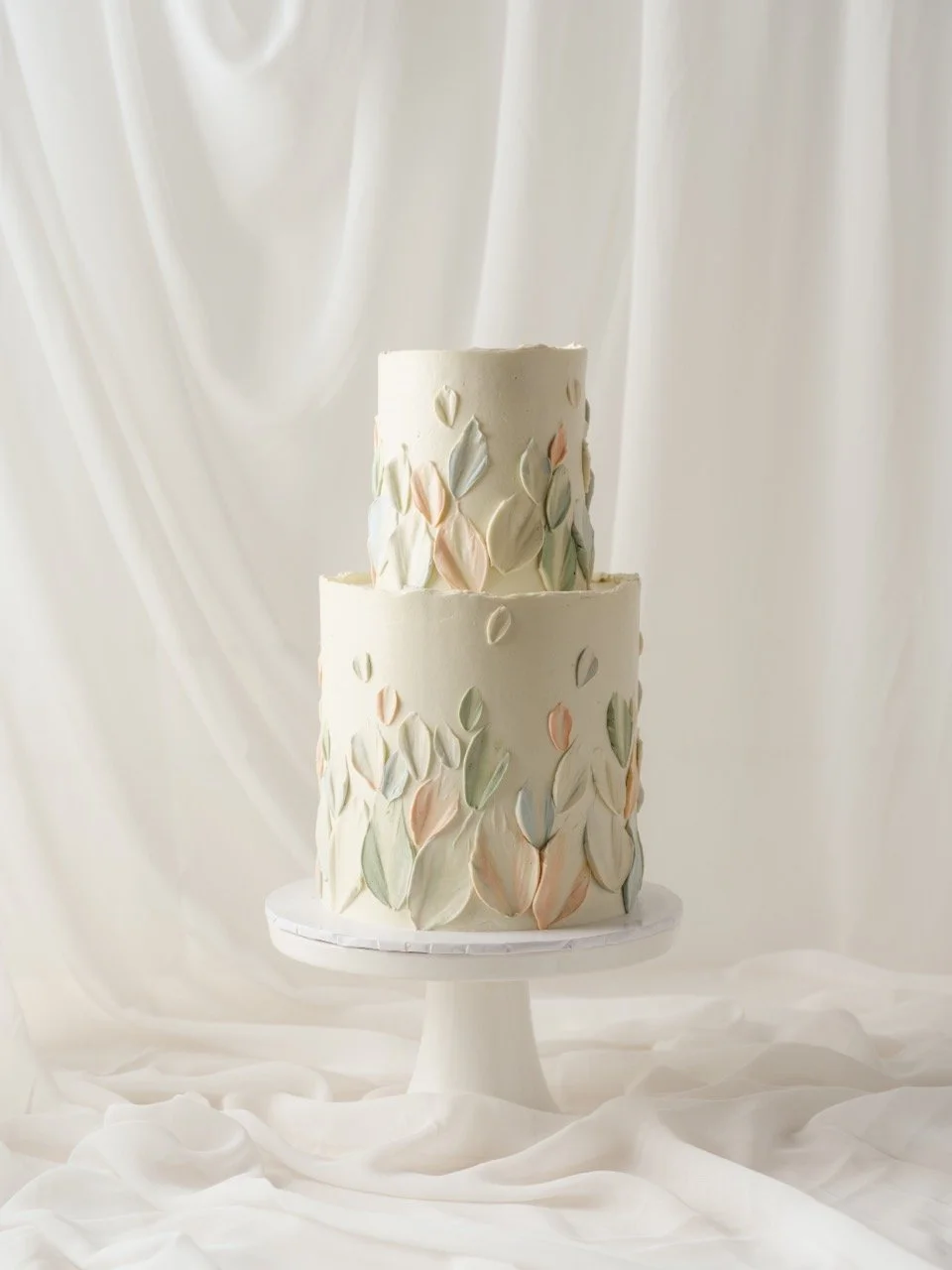 Two-tiered white wedding cake decorated with pastel tulip petal buttercream textures, displayed on a white cake stand against a white draped fabric background.