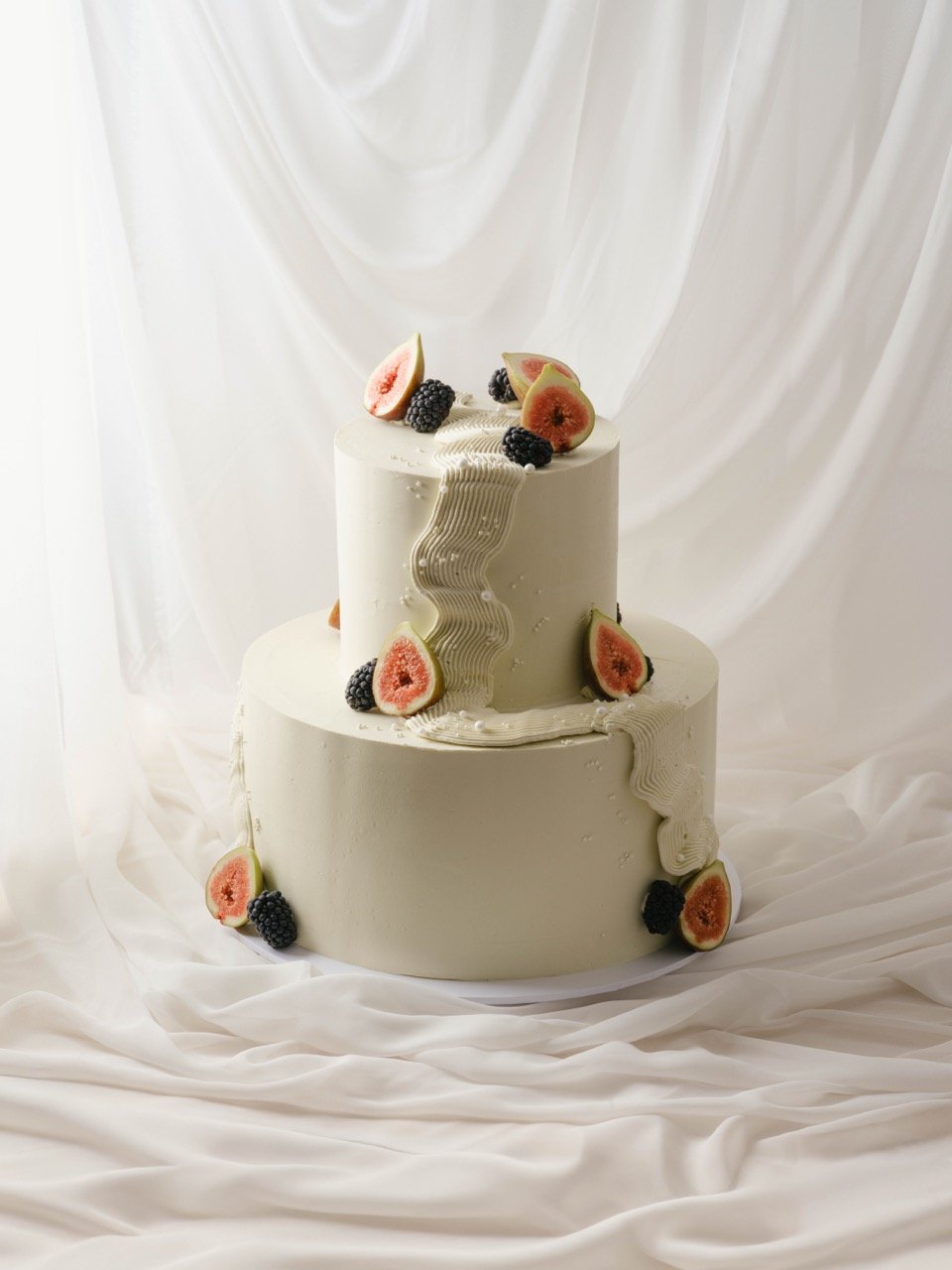 Two-tiered white cake decorated with slices of fig and blackberries, with white icing piped down the sides and pearl sprinkles, set against a white fabric backdrop.