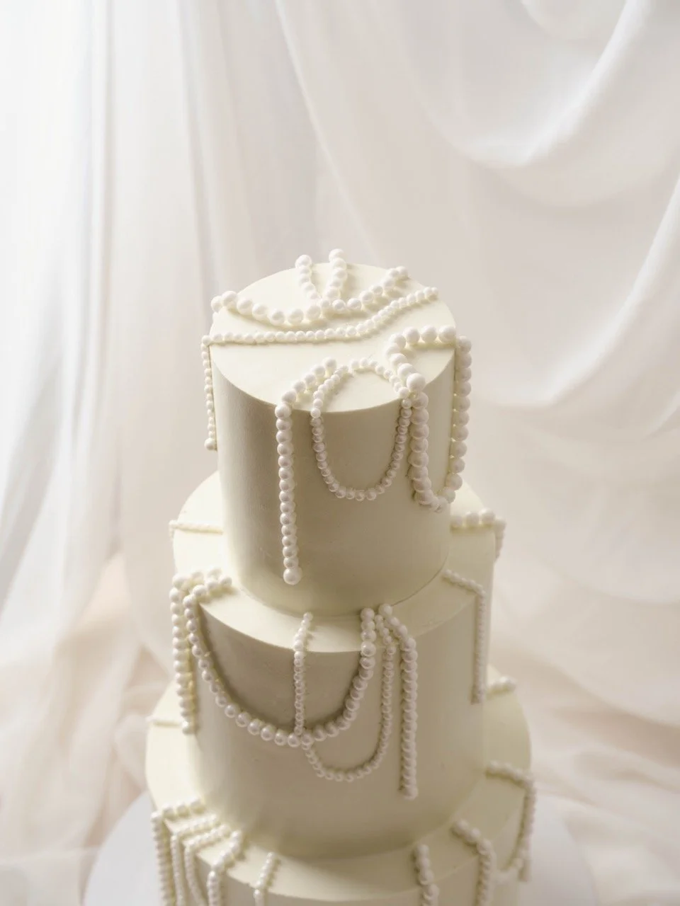 Three-tiered white wedding cake decorated with pearl strings, set against a soft white fabric background.