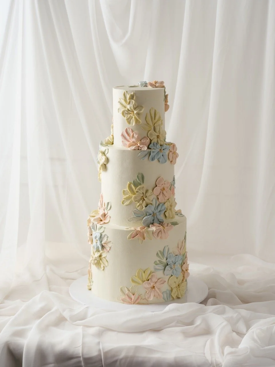 A tall, three-tiered wedding cake decorated with pastel piped buttercream flowers on a white cloth background.