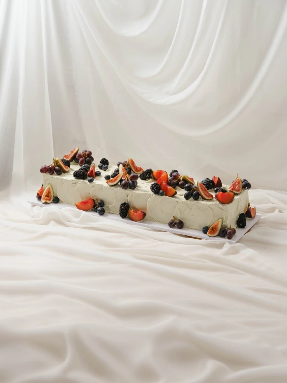 A long rectangular wedding cake decorated with assorted fresh fruits including strawberries, blackberries, blueberries, grapes, and figs, set against a white satin background.