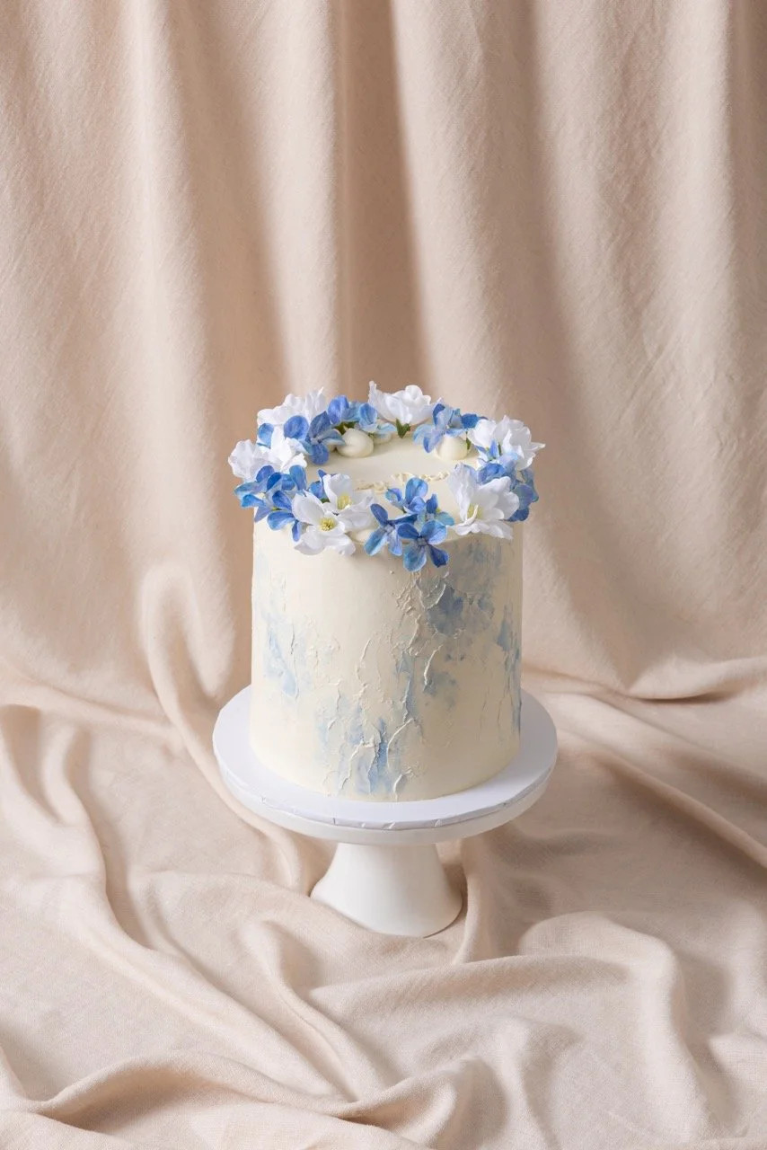 A white birthday cake decorated with blue and white flowers on top, placed on a white cake stand against a beige curtain background.