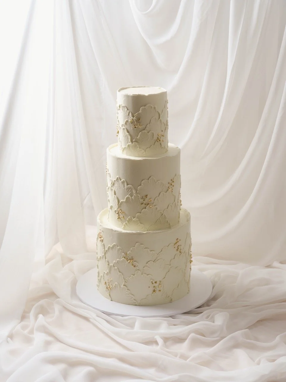 Three-tiered white wedding cake with fine line lace buttercream texture design and gold accents, set against white draped fabric.
