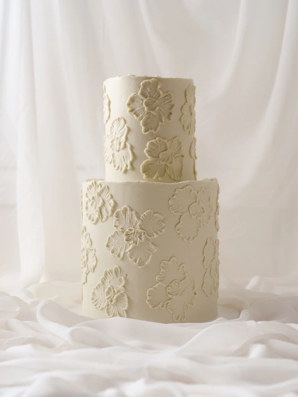 Two-tiered white wedding cake decorated with textured flower patterns, set against sheer white fabric background.