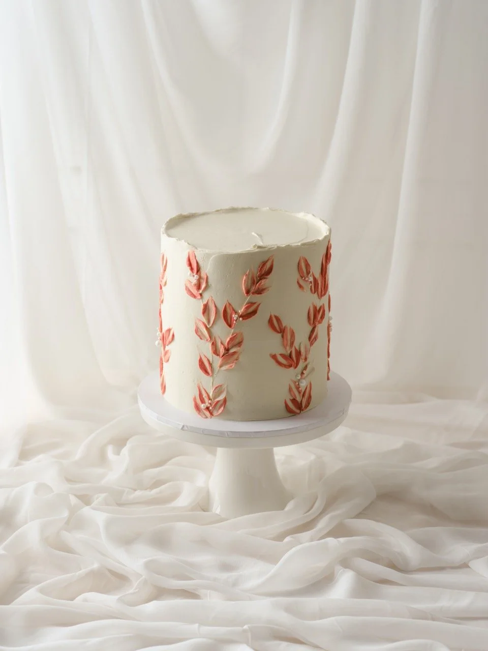 A white cake decorated with pink and red leaf buttercream texture patterns, placed on a white cake stand against a white draped fabric background.