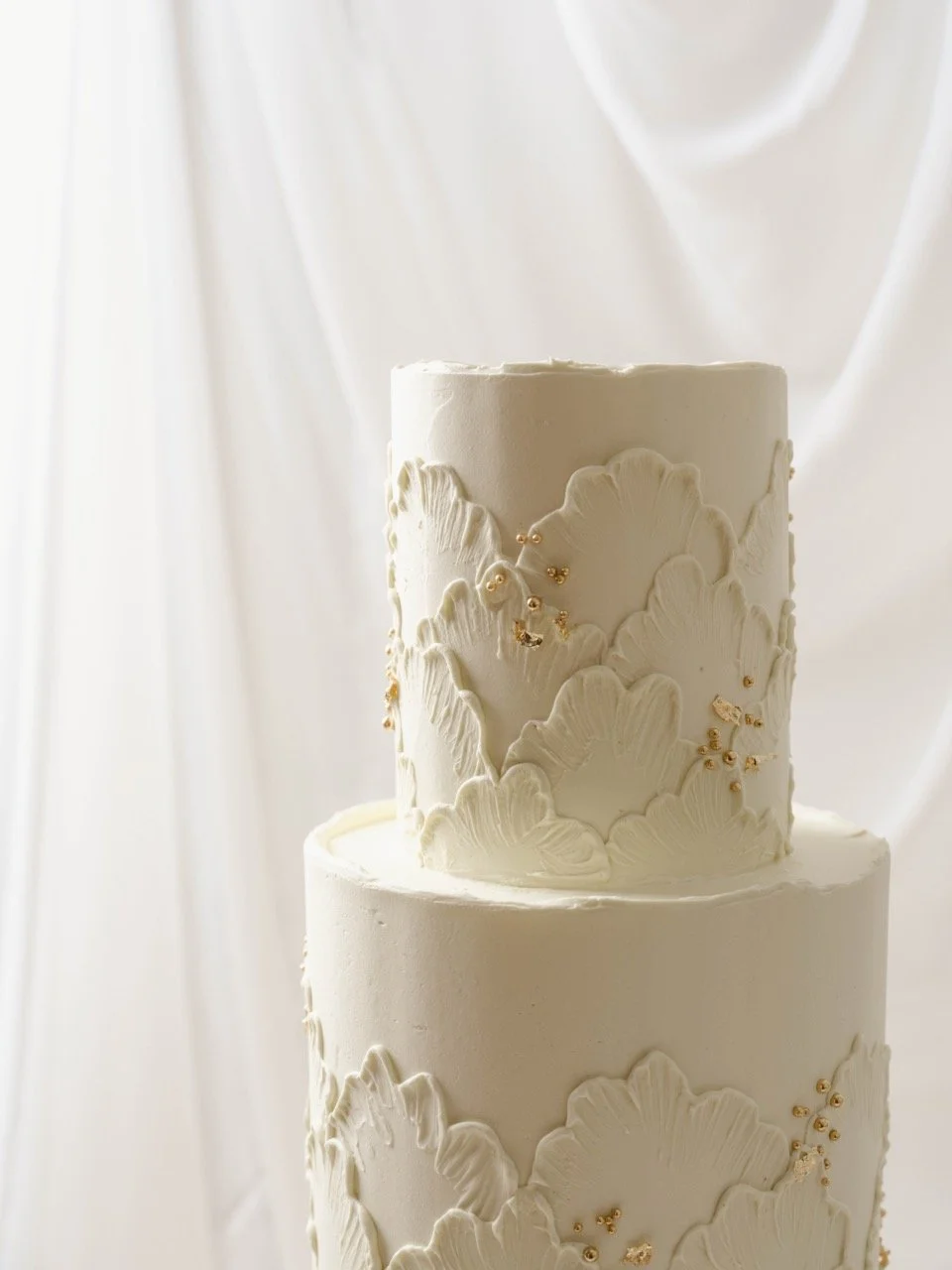 Three-tiered white wedding cake with fine line lace buttercream texture design and gold accents, set against white draped fabric.