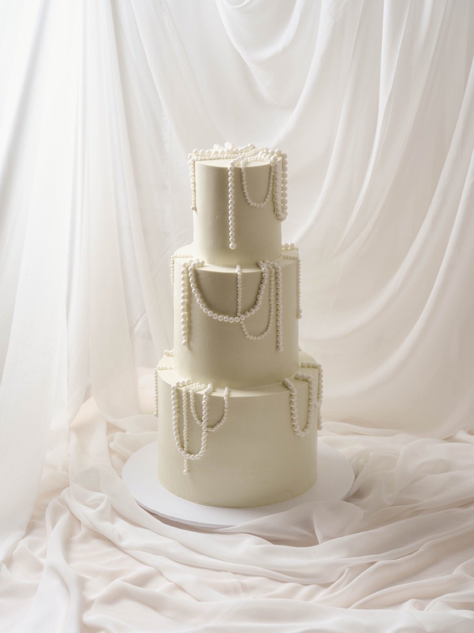 Three-tiered white wedding cake decorated with pearl strings, set against a soft white fabric background.