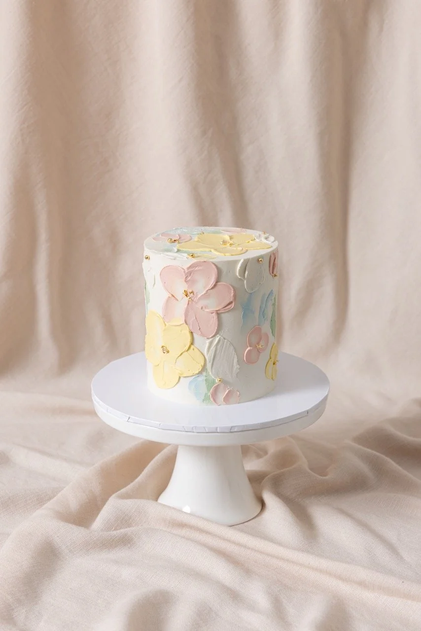 A small, round cake with pastel-colored floral decorations on a white pedestal stand, set against a beige fabric backdrop.