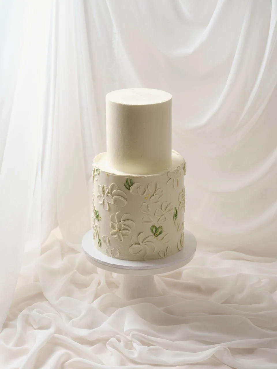 A two-tier white wedding cake with floral buttercream textures and soft green on a white cake stand, set against a draped white fabric background.
