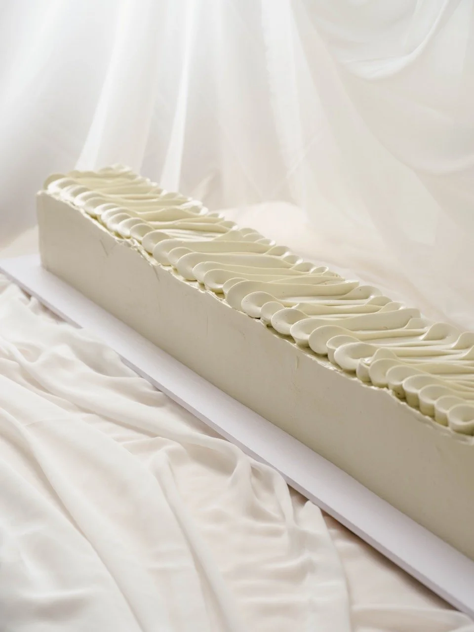 A long rectangular white slab cake with minimalist piping on top, displayed on a table with white satin fabric and white drapery in the background.