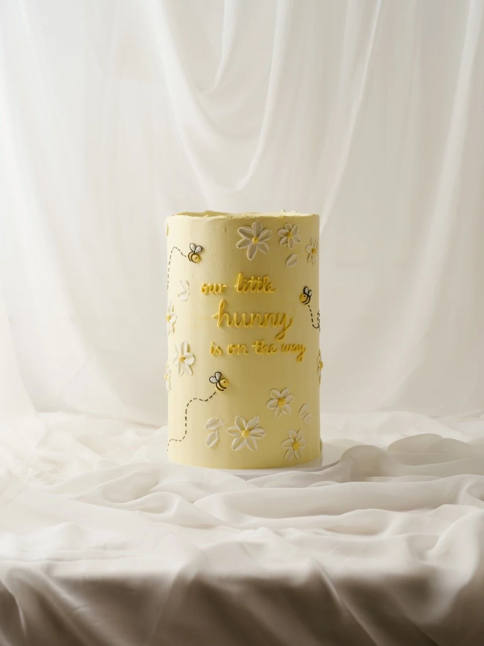A tall, single tier cake decorated with white flowers, small bees, and handwritten-style yellow text that reads 'our little hunny is on the way,' placed on a white, crinkled fabric background with draped white fabric backdrop.