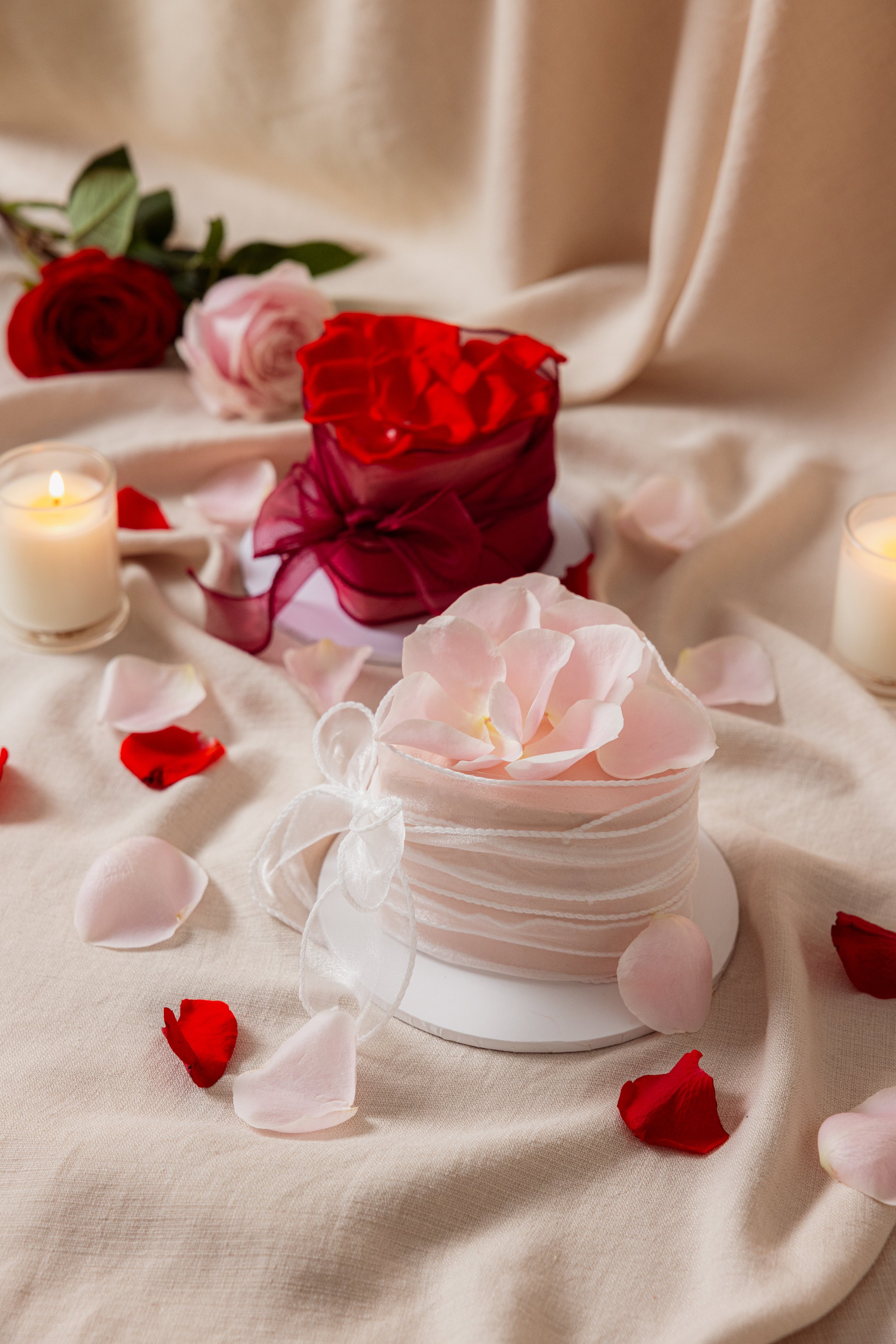 A romantic setup with a cake decorated with pink petals, scattered rose petals, candles, and roses on a cream-colored cloth background.