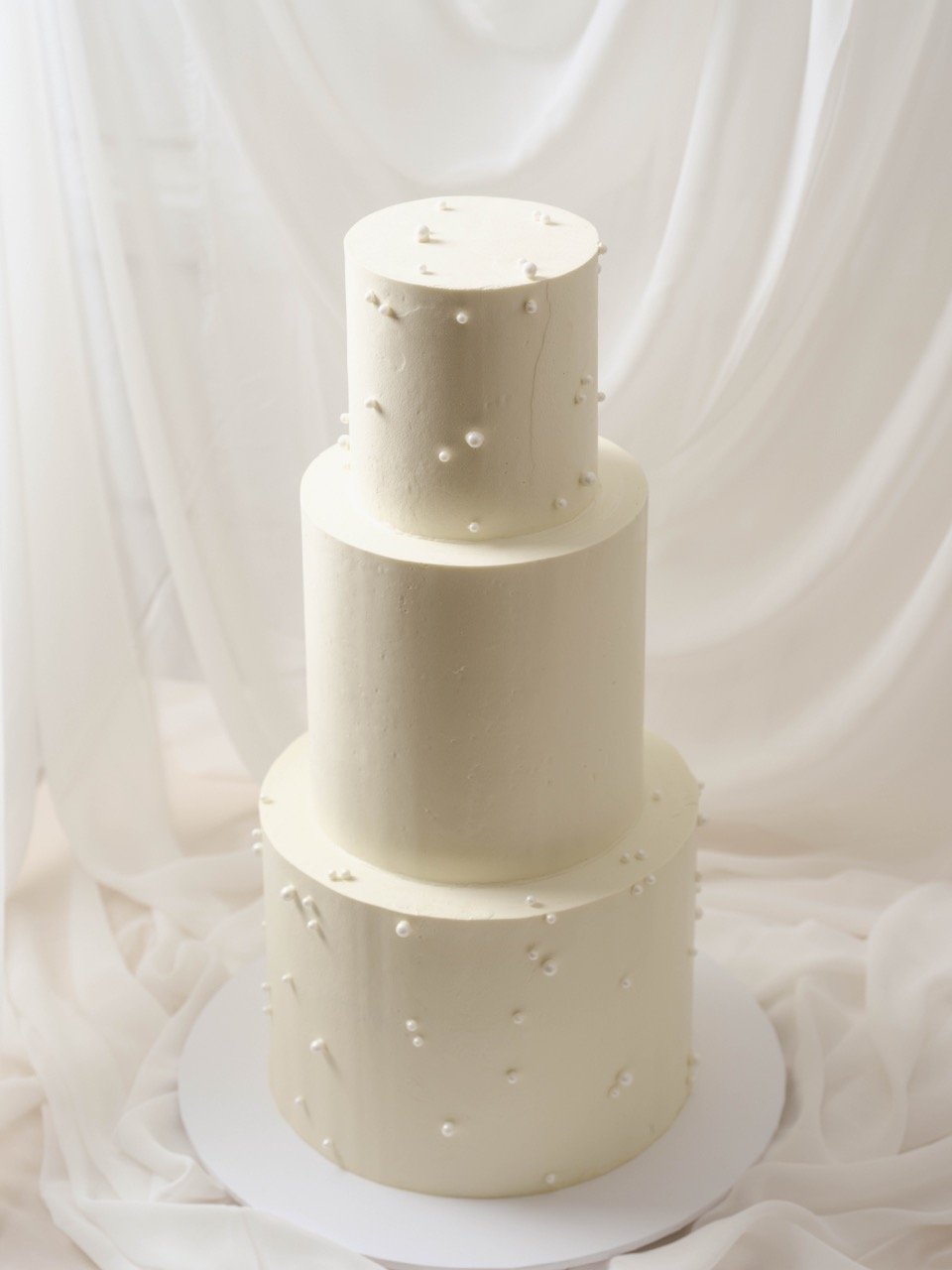 Three white tiered cake with minimalist pearl sprinkles, set against light, draped fabric background.