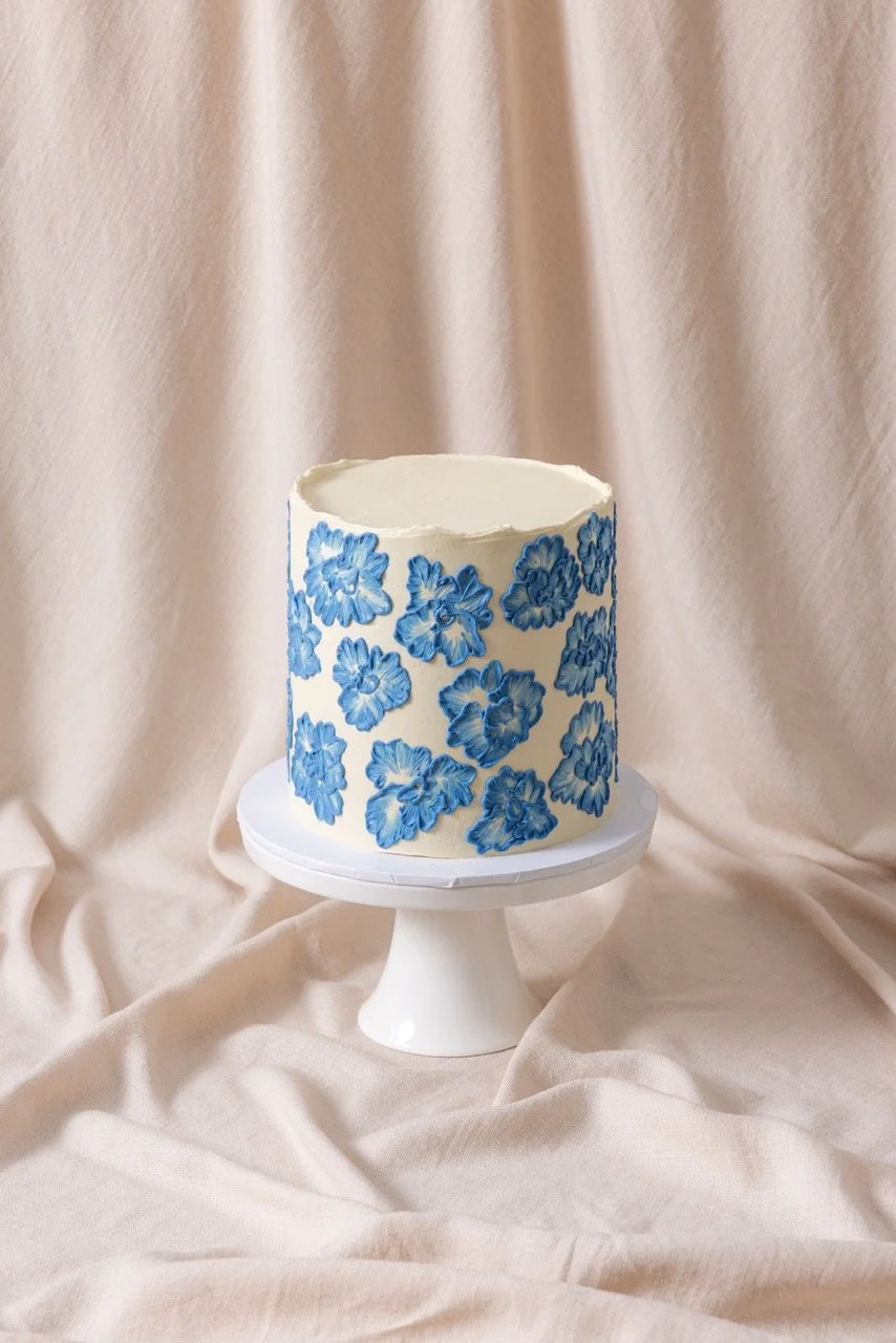 A white cake decorated with blue floral designs on a white cake stand against a beige fabric backdrop.