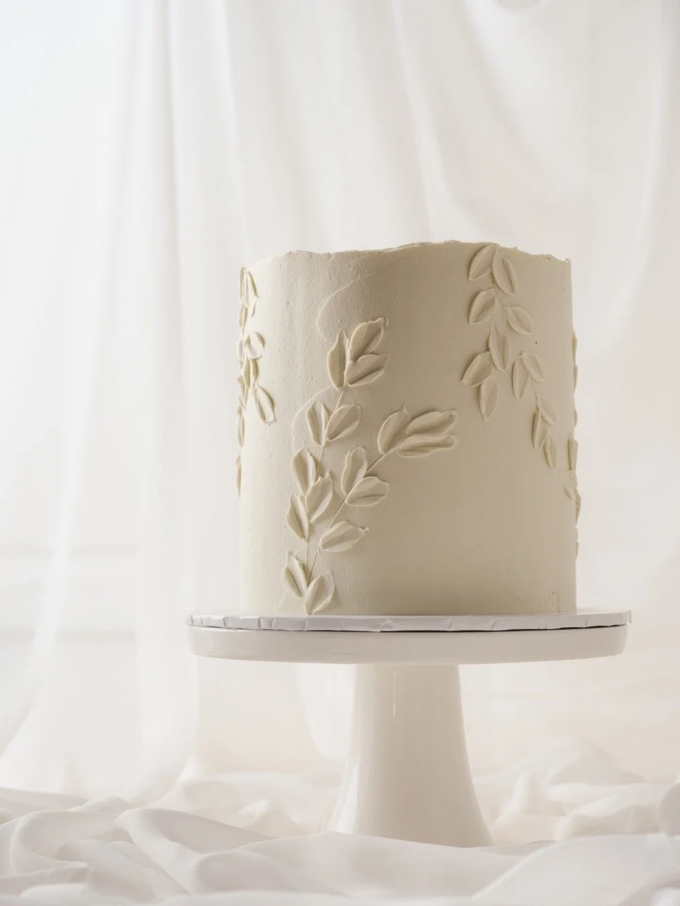 A white cake with a leaf textured buttercream design on a white cake stand, with a soft white background.