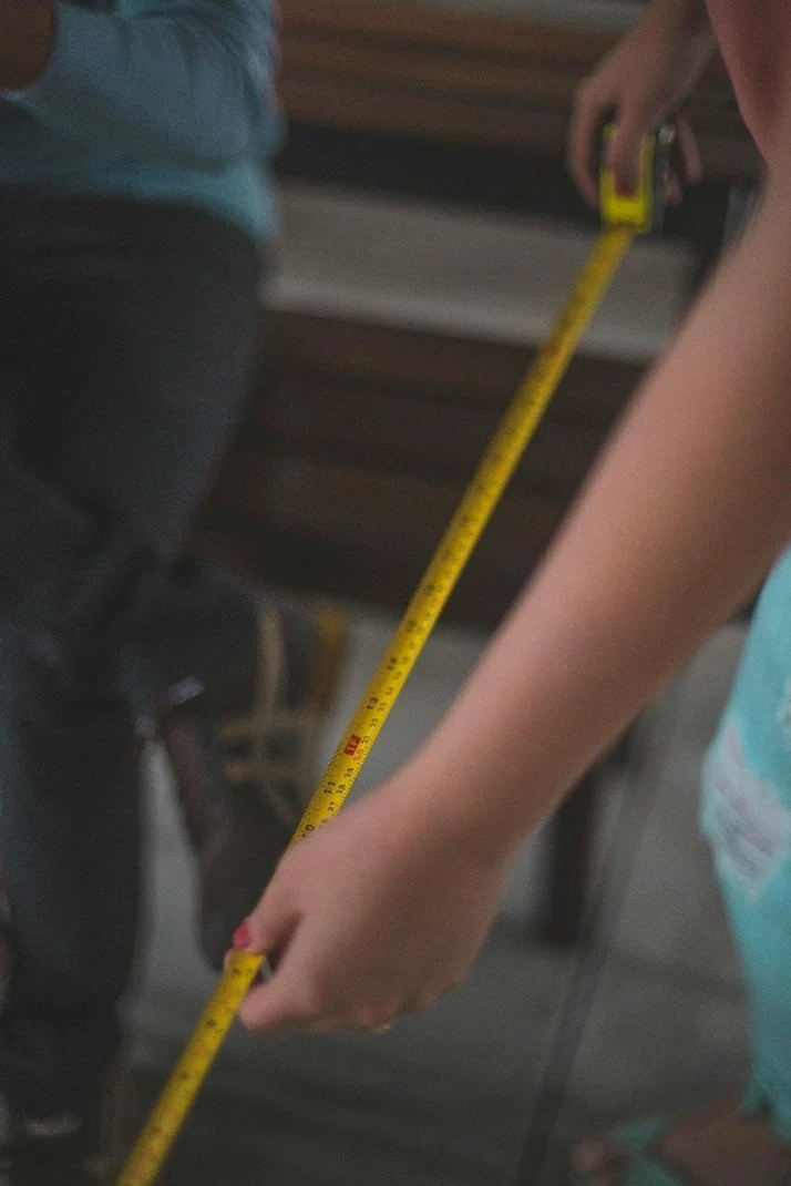A person's arm holding a yellow measuring tape, measuring a distance. Part of a person in a turquoise shirt and another person in a blue shirt are also visible.