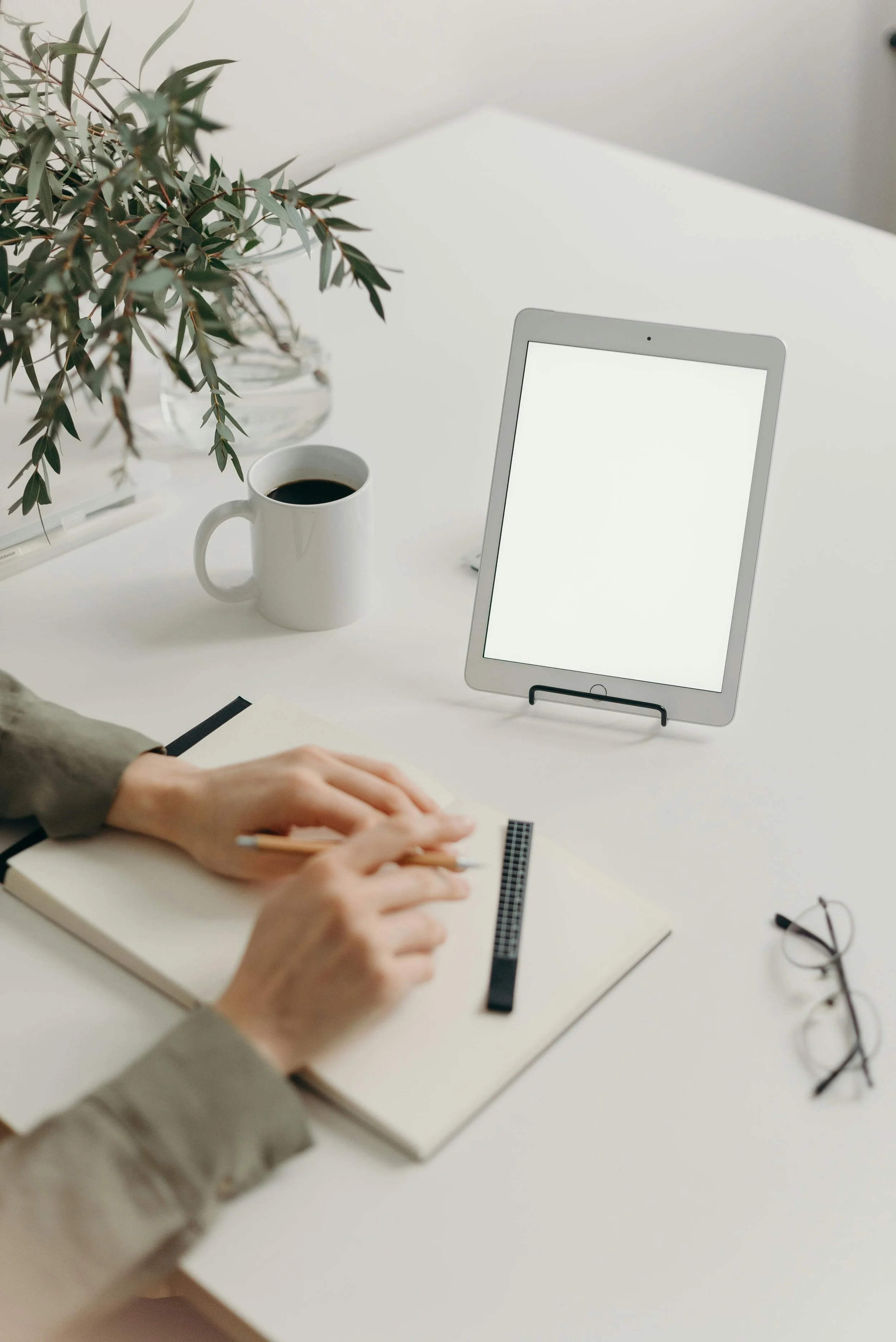 A person's hands writing in a notebook with a pencil, a ruler, and a pair of glasses on a white desk. On the desk are a tablet, a mug of black coffee, a plant in a glass bowl, and a laptop.