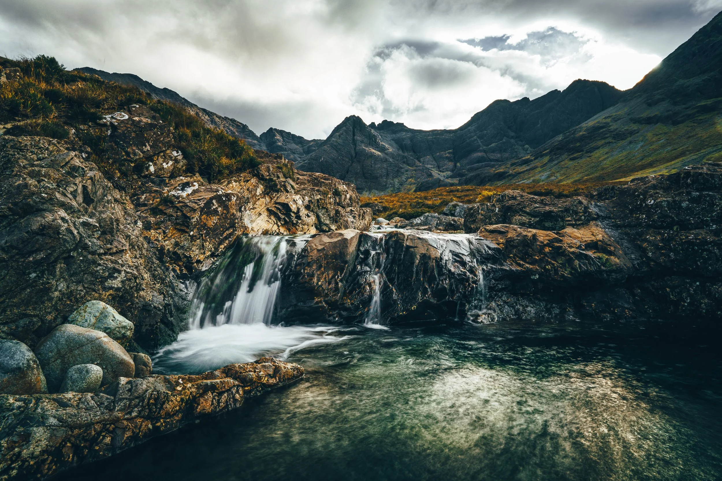 A mountain stream flowing over rocks with mountains and cloudy sky in the background.