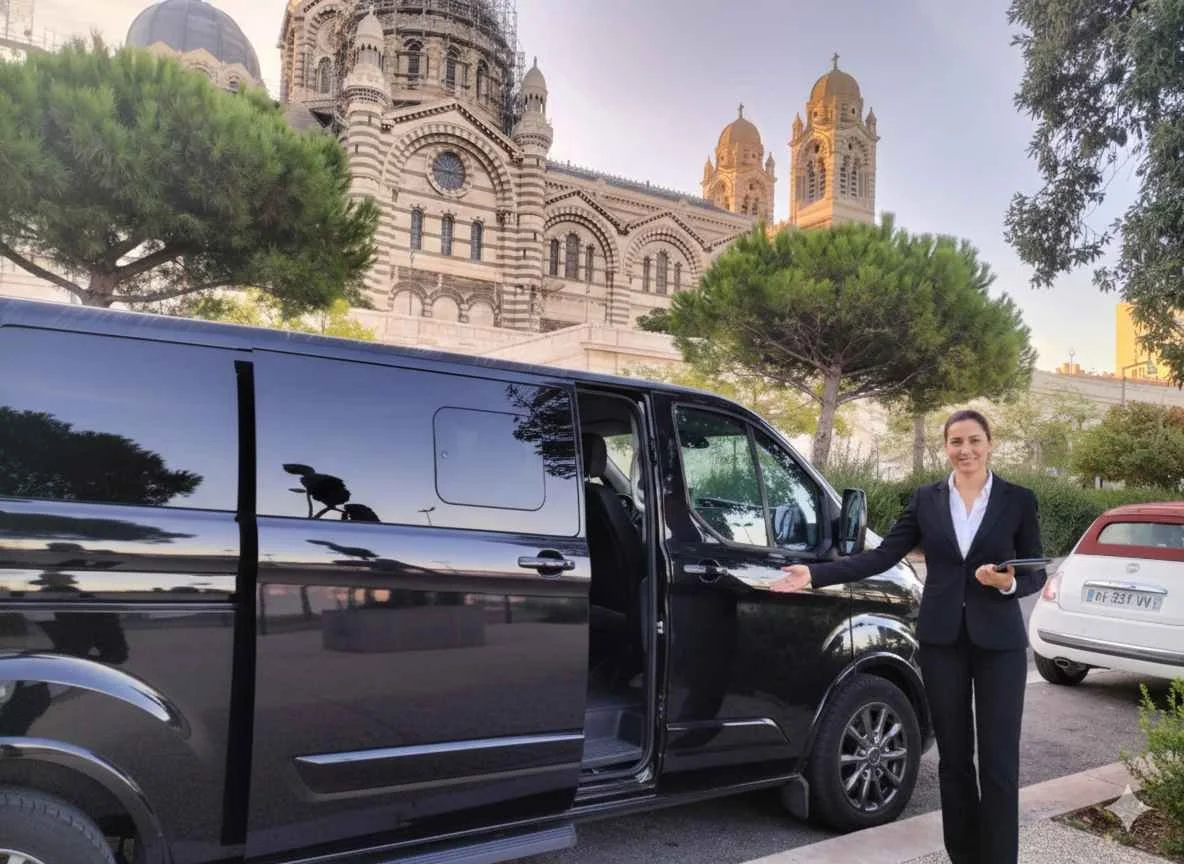 A woman in a business suit gesturing towards a black van parked on the street, with the basilica of Notre-Dame de la Garde visible in the background, in Marseille, France.