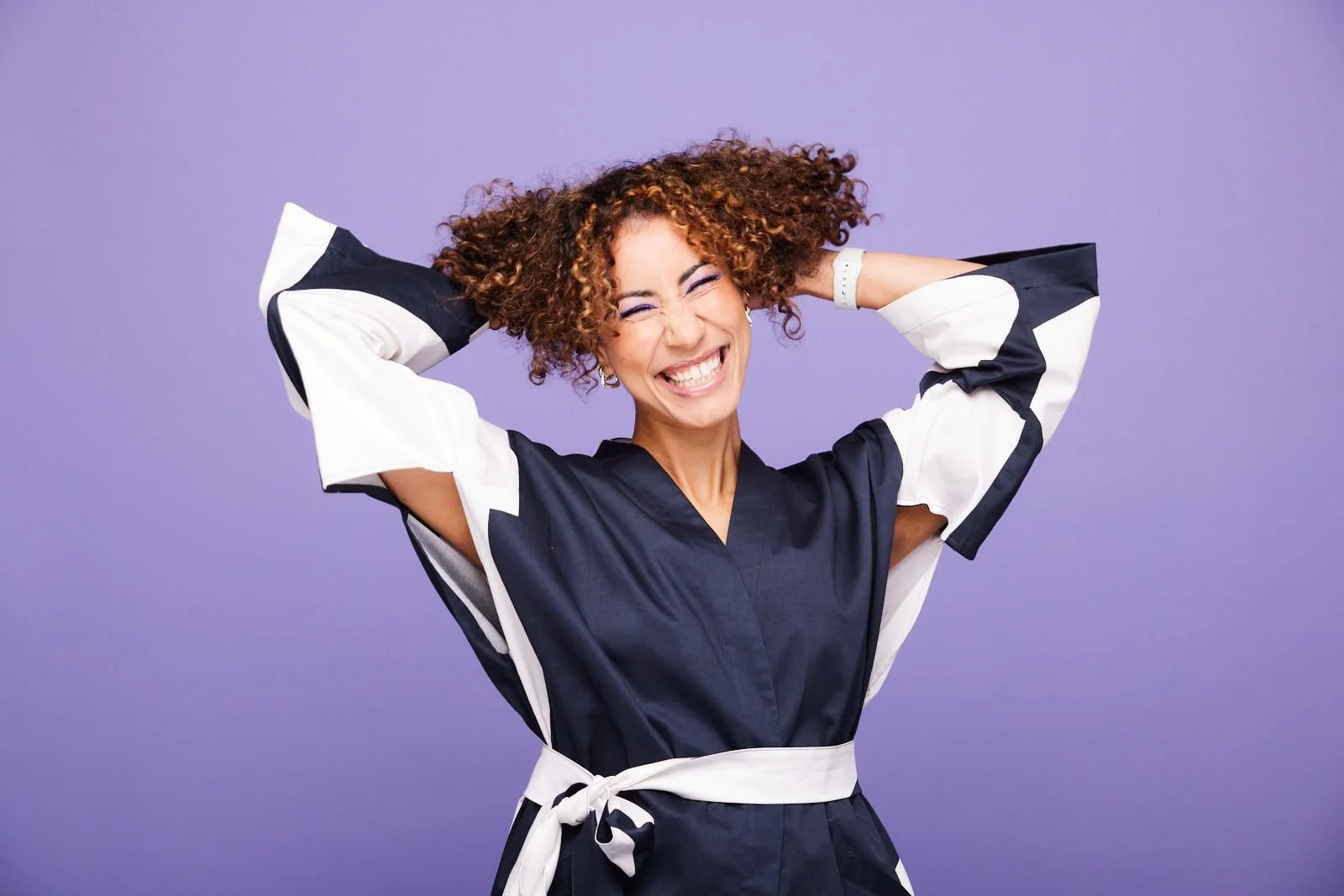 A woman with curly hair wearing a black and white dress smiling with her eyes closed and holding her hands behind her head, standing against a purple background.