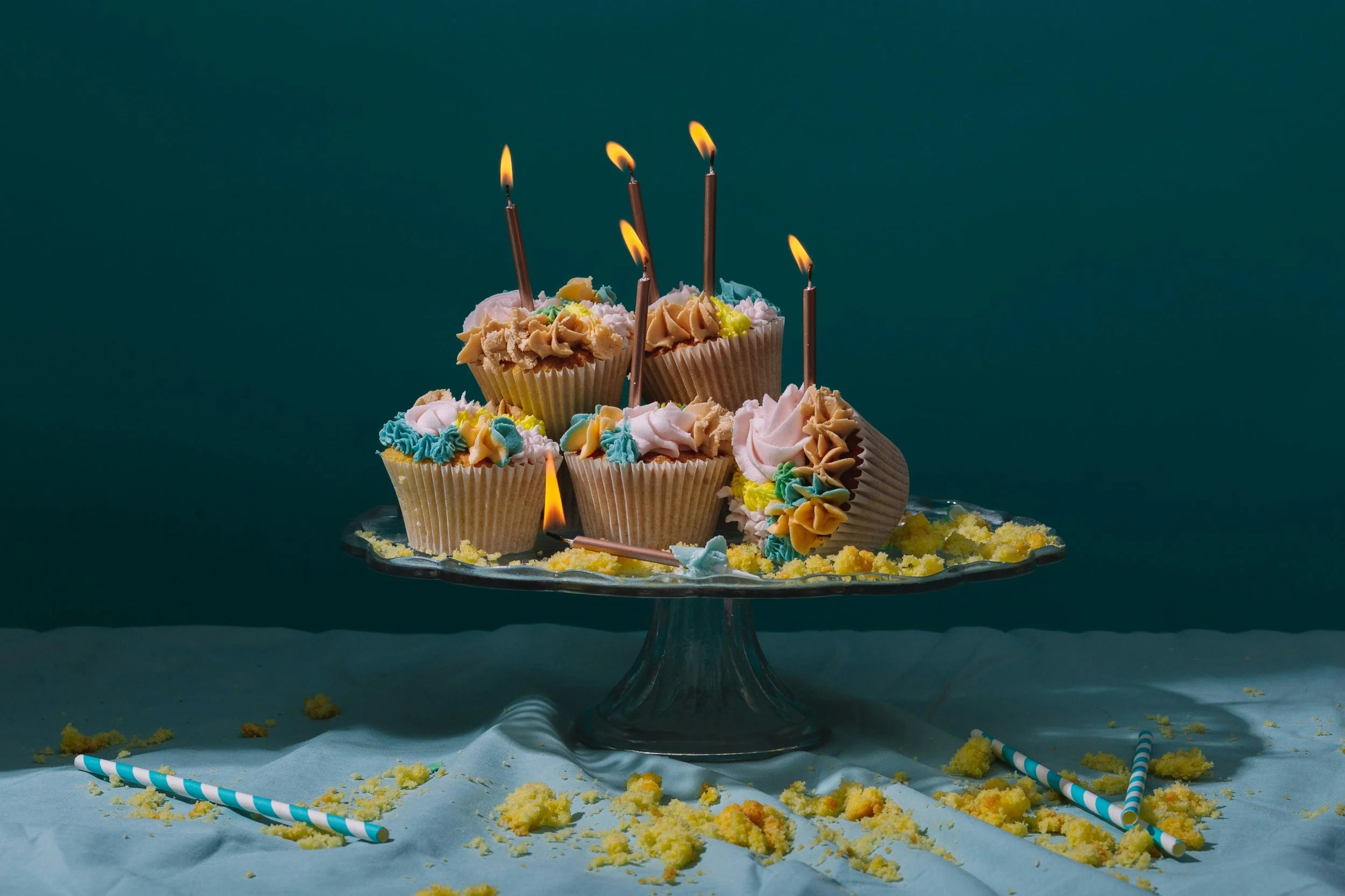 A birthday cake decorated with colorful frosting and topped with lit birthday candles. The cake is on a glass cake stand, with birthday candles and cake crumbs scattered around on the table.