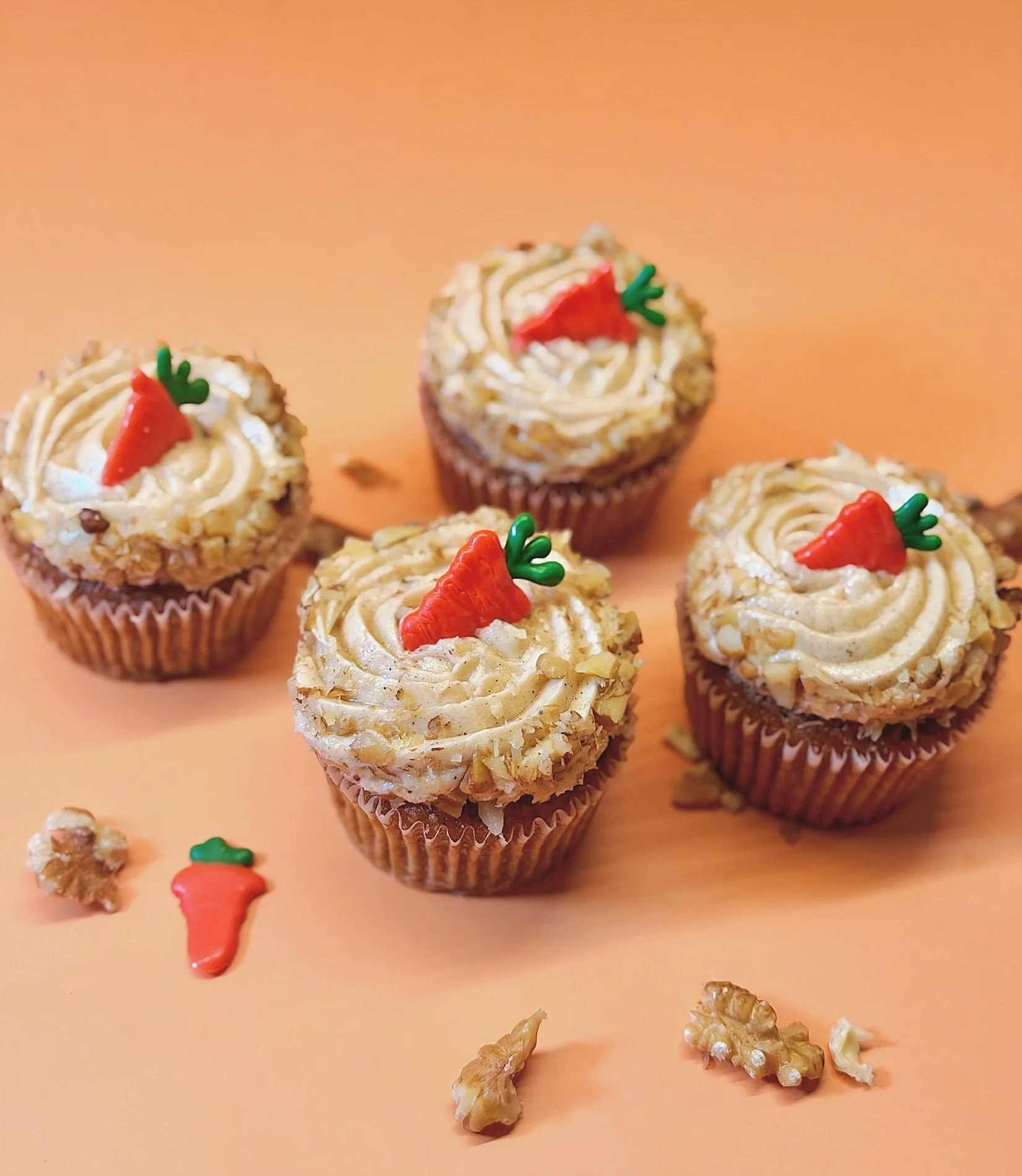 Cupcakes decorated with frosting and carrot-shaped toppers, with scattered walnuts and small food decorations on an orange background.