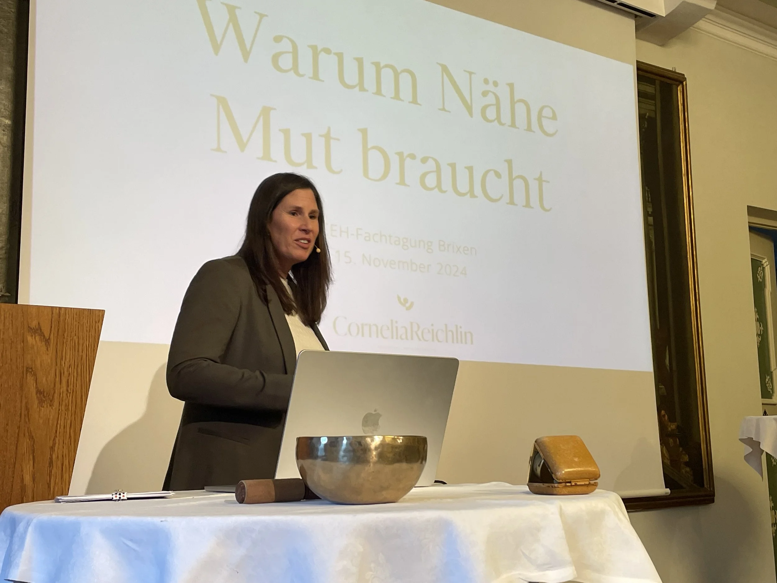 Eine Frau steht hinter einem Rednerpult bei einer Präsentation. Im Hintergrund ist eine Leinwand mit Text zu sehen, darunter der Name Cornelia Reichlin. Auf dem Tisch vor ihr stehen ein gusseiserner Topf, ein Holzblock, ein fotokopiertes Dokument, eine kleine Ledertasche und ein Holzhammer.