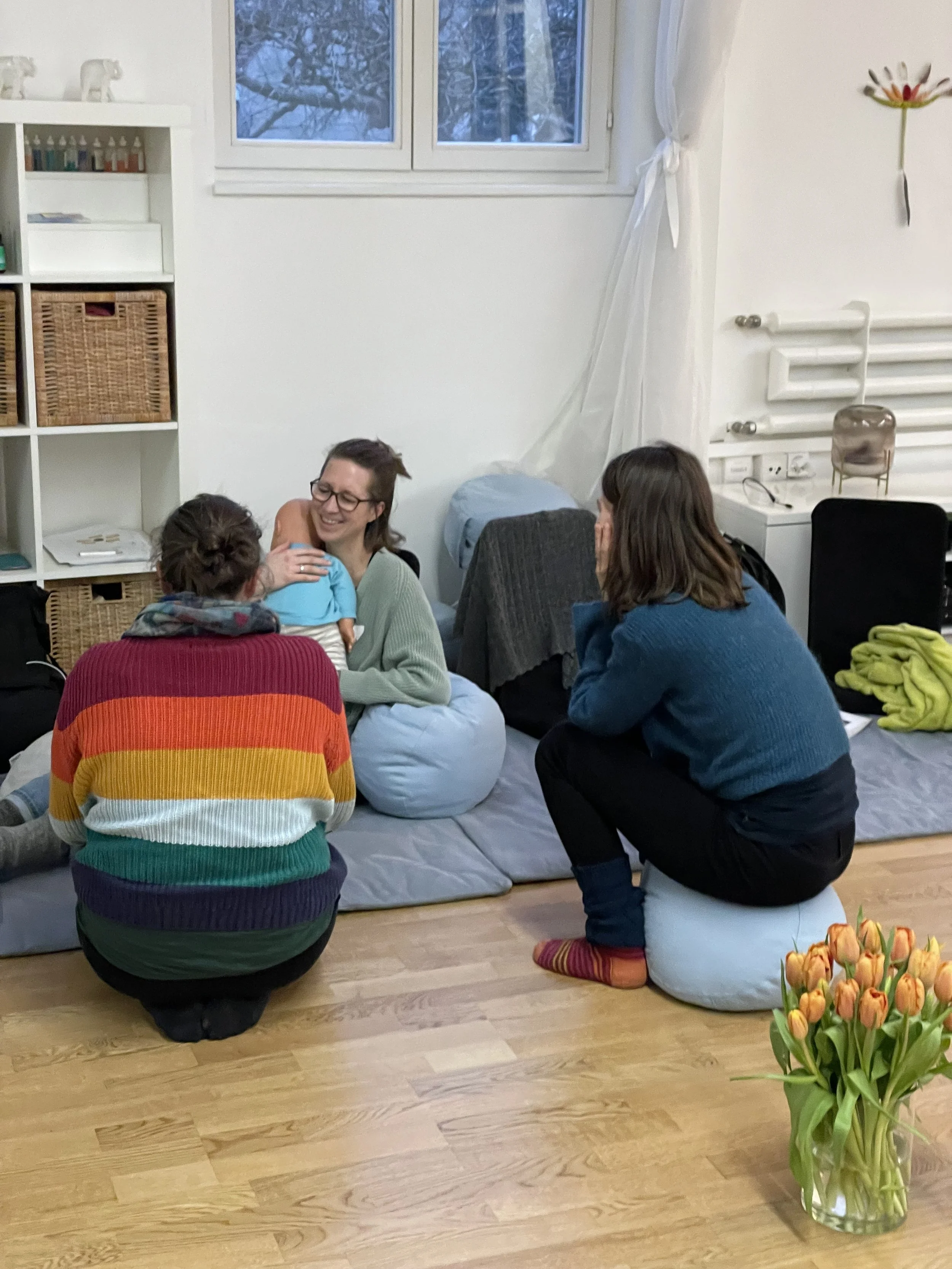 Vier Frauen sitzen und stehen in einem Wohnzimmer. Sie lachen und unterhalten sich. Es gibt eine Vase mit orangefarbenen Tulpen im Vordergrund, eine weiße Wand mit Fenster im Hintergrund, und eine weiße Heizung an der Wand.