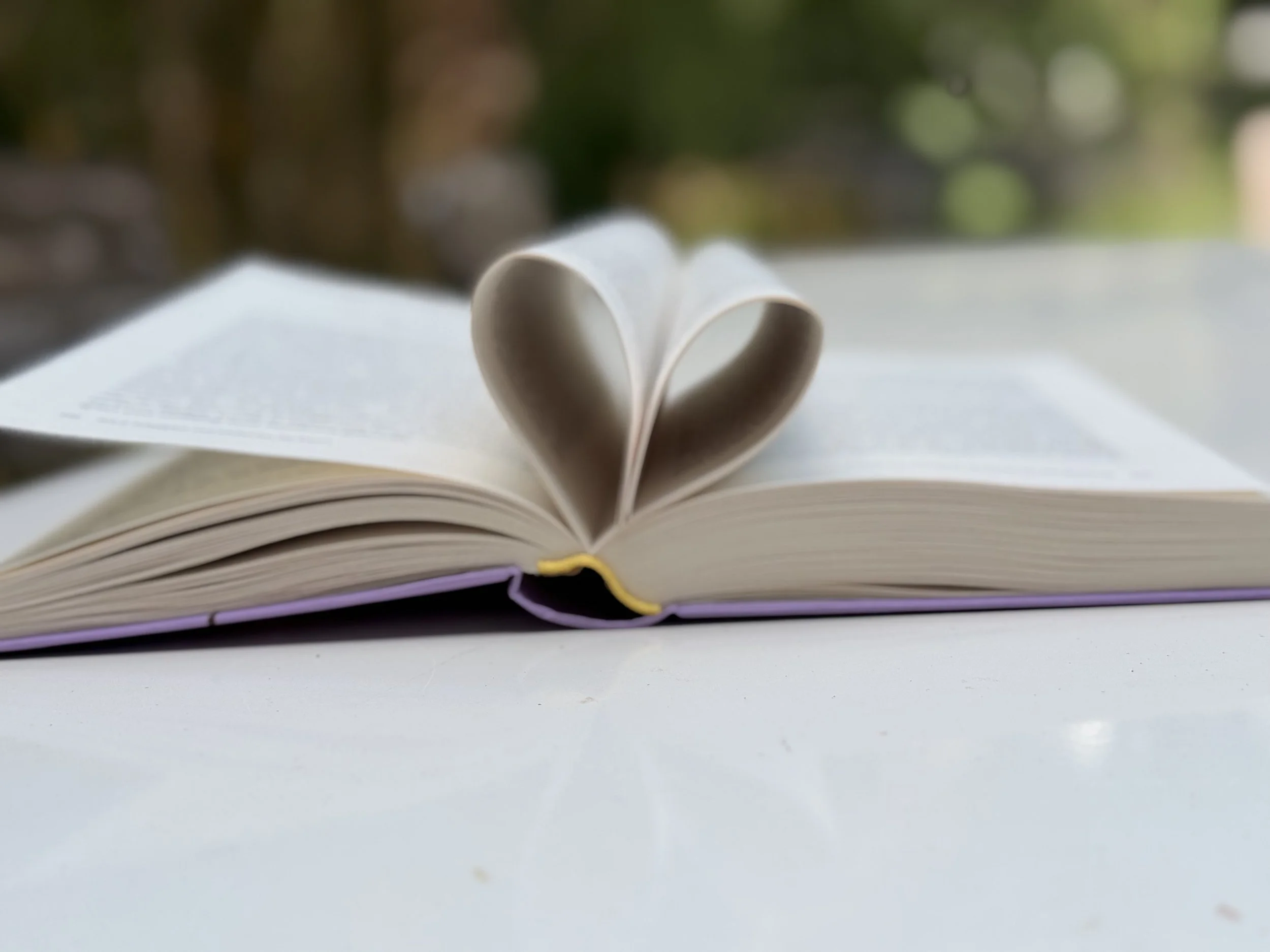 Offenes Buch mit Herz auf den Seiten, auf einem weißen Tisch, mit unscharfem grünen Hintergrund.