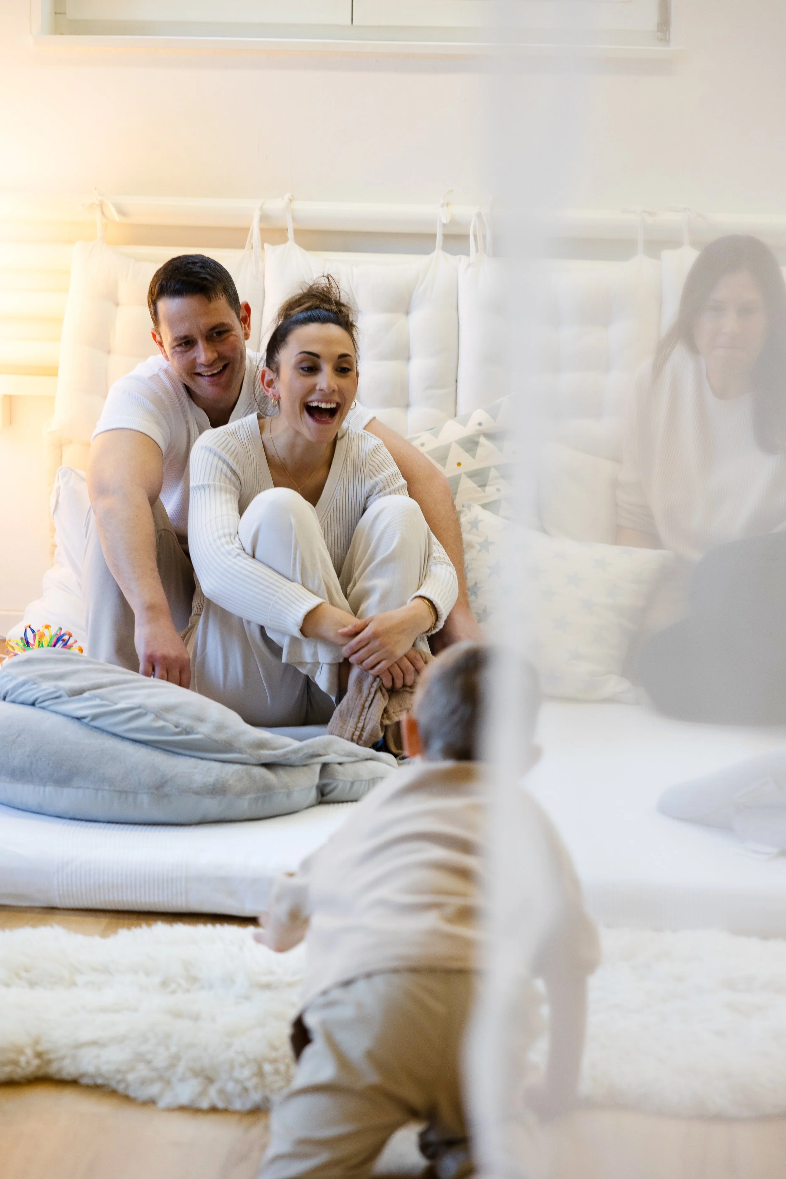 Ein Mann und eine Frau sitzen auf einem Bett und lachen miteinander, während sie ein kleines Kind, das in weißen Kleidern gekleidet ist, ansehen. Im Hintergrund sind weiße Kissen und eine Kopfstütze. Es scheint ein angenehmer und fröhlicher Moment im Schlafzimmer zu sein.