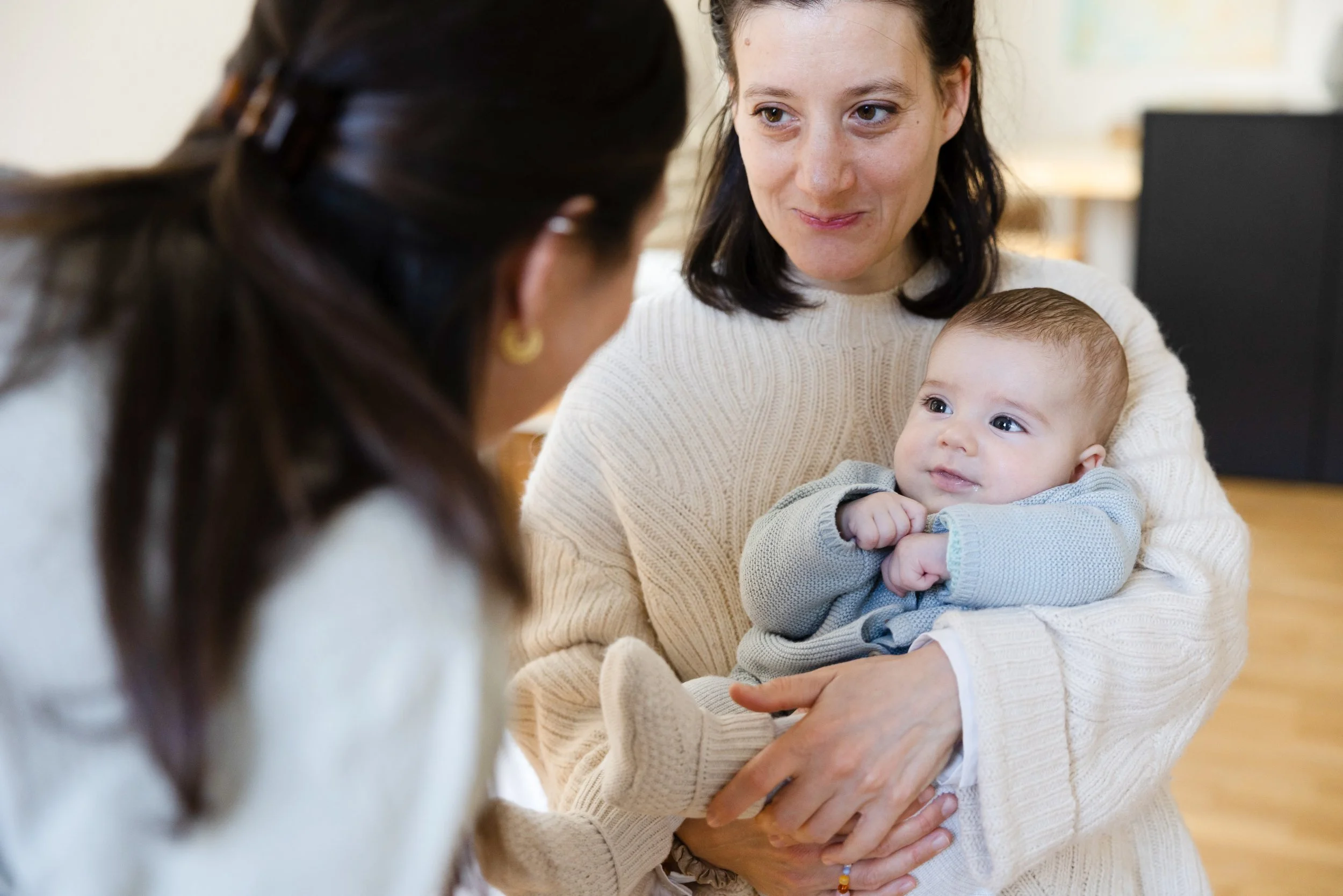 Eine Frau hält ein Baby im Arm, während eine andere Person, nur teilweise sichtbar, im Vordergrund ist.