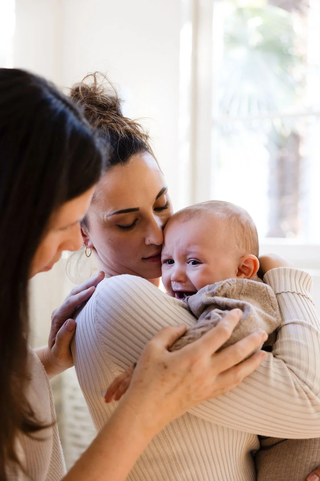 Zwei Frauen halten ein weinendes Baby in einem Raum mit natürlichem Licht.