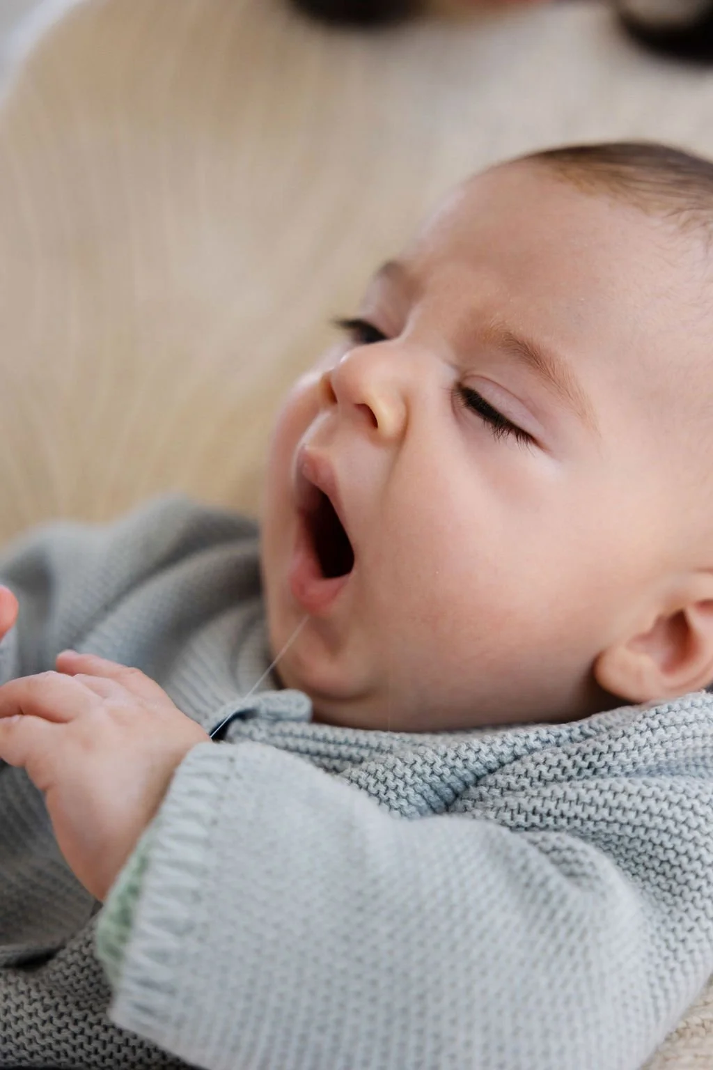 Ein Baby, das gelangweilt oder müde ist, mit offenem Mund und geschlossenen Augen, während es auf einer weichen Oberfläche liegt, getragen von einer Person.